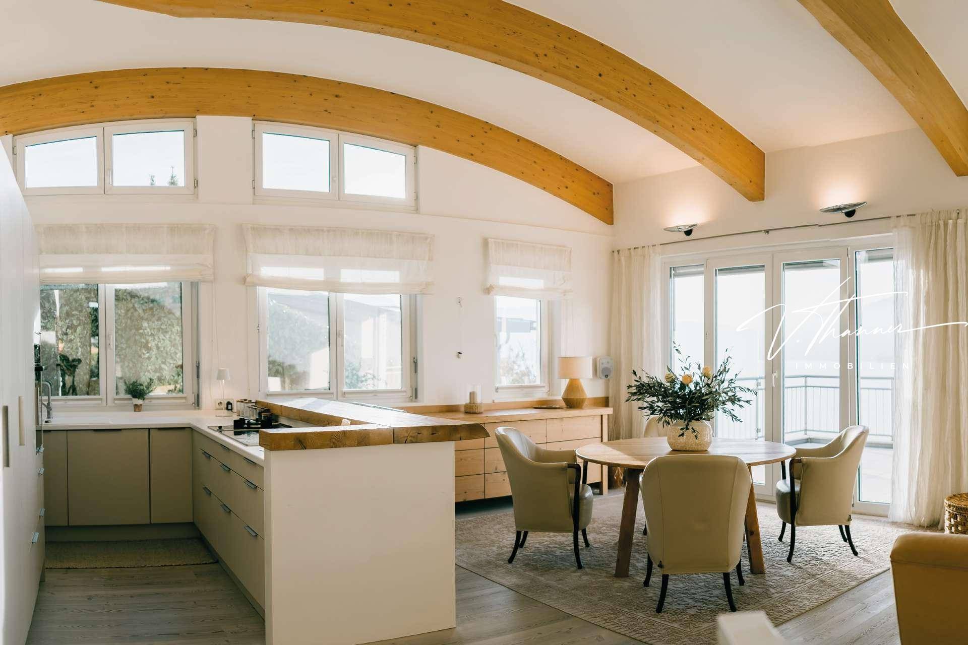Heller offener Wohn-Essbereich mit Holzbalken, moderner Küche und Seeblick durch große Fenster.