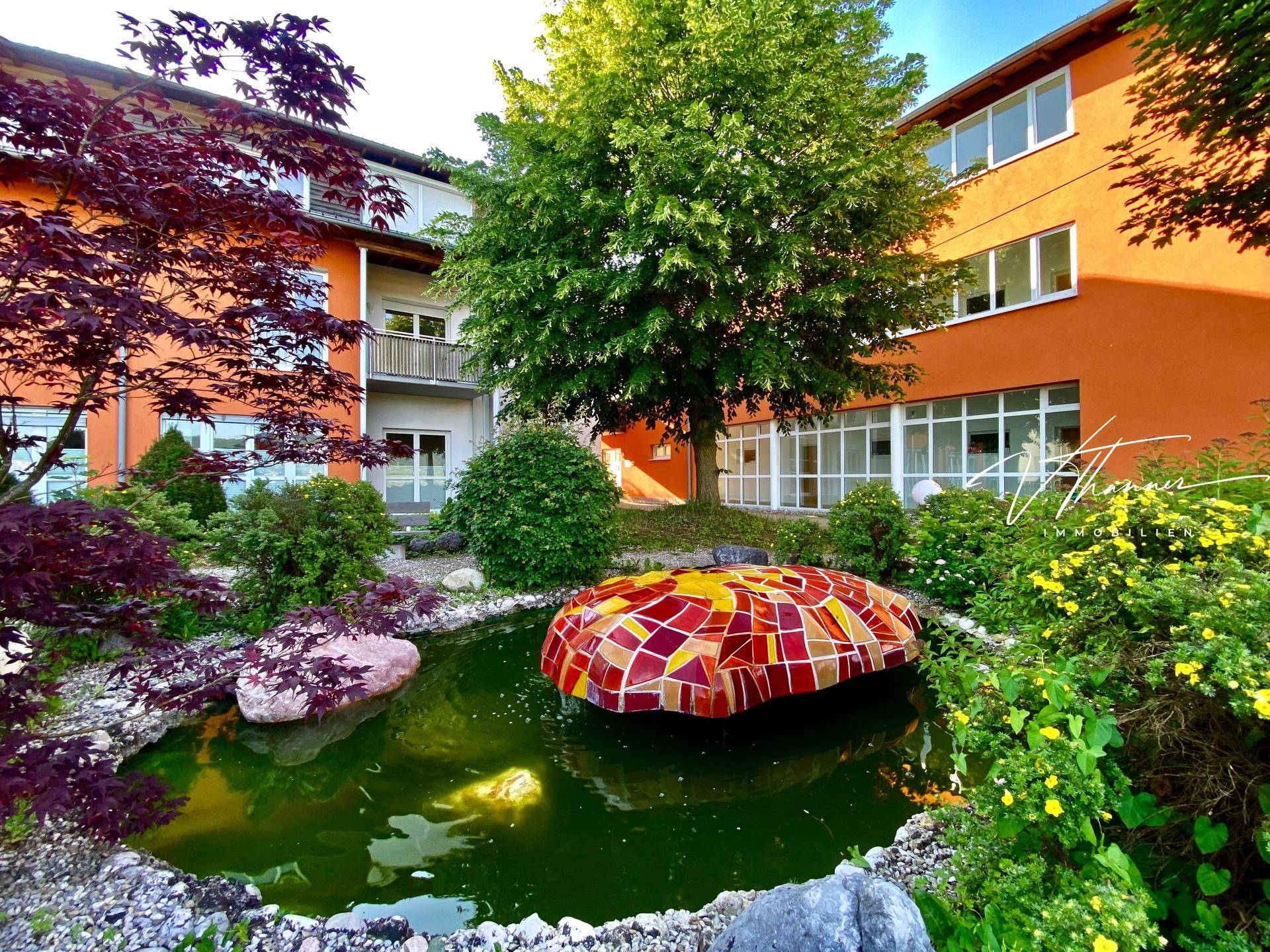 Malerischer Gartenbereich mit einem Teich, einer farbenfrohen Mosaikskulptur und üppiger Bepflanzung vor einem Gebäude.
