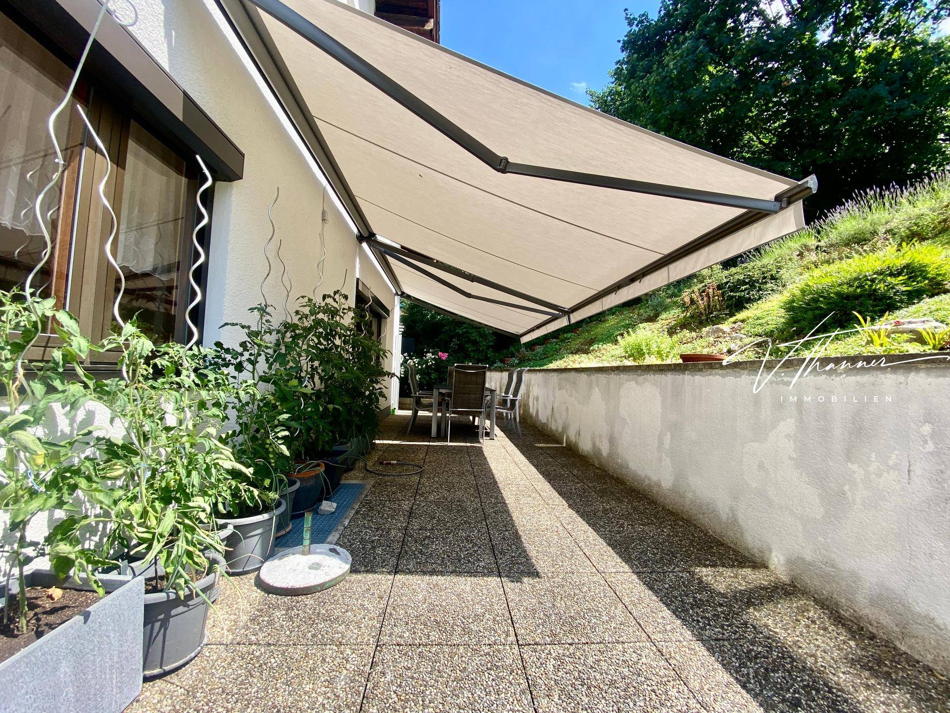Überdachte Terrasse mit ausgefahrener Markise, Gartenmöbeln und Pflanzen in Töpfen.