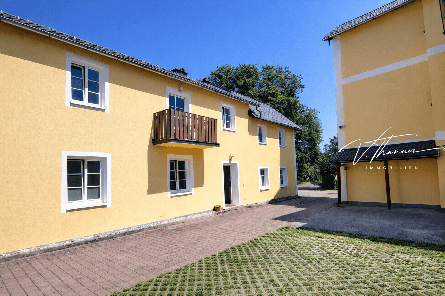 Gelbe Gebäudefassade mit weißen Fenstern, kleinem Balkon und gepflastertem Vorplatz.