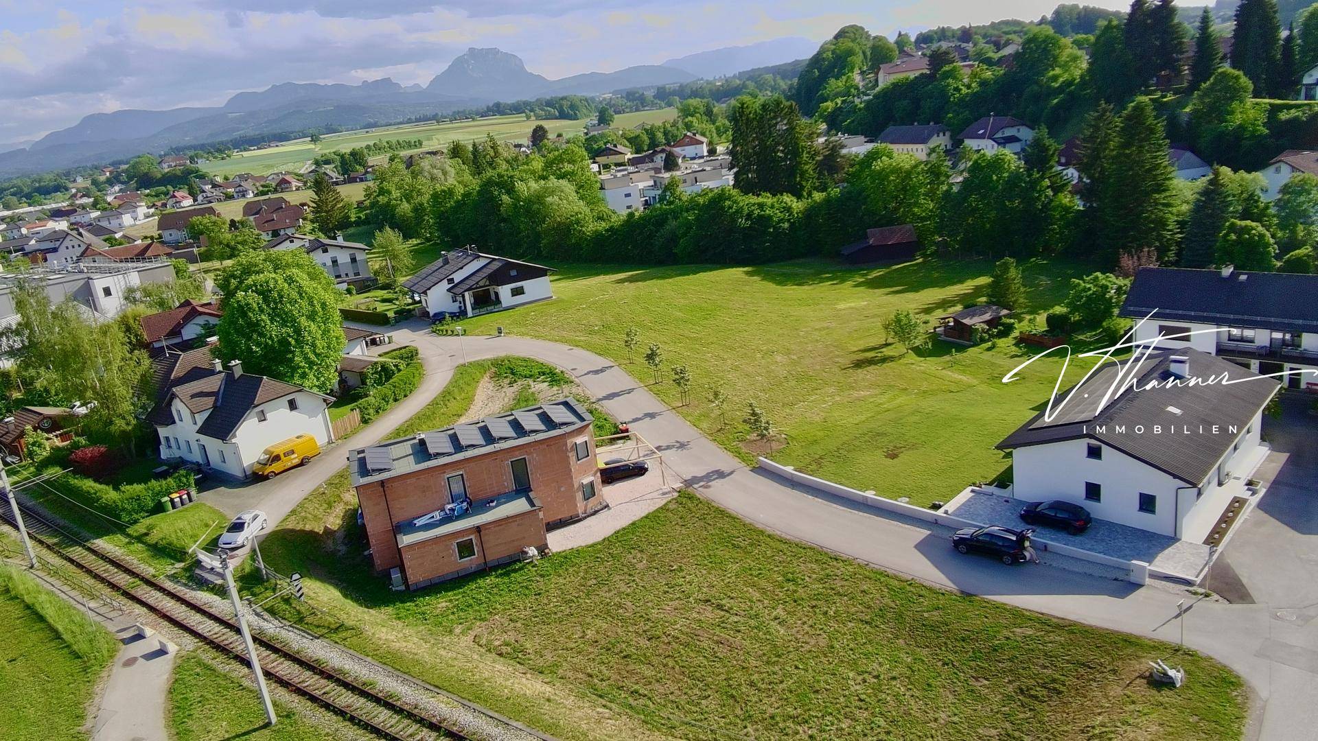 Luftaufnahme einer Wohnsiedlung mit Häusern, viel Grün und Bergen im Hintergrund unter blauem Himmel.