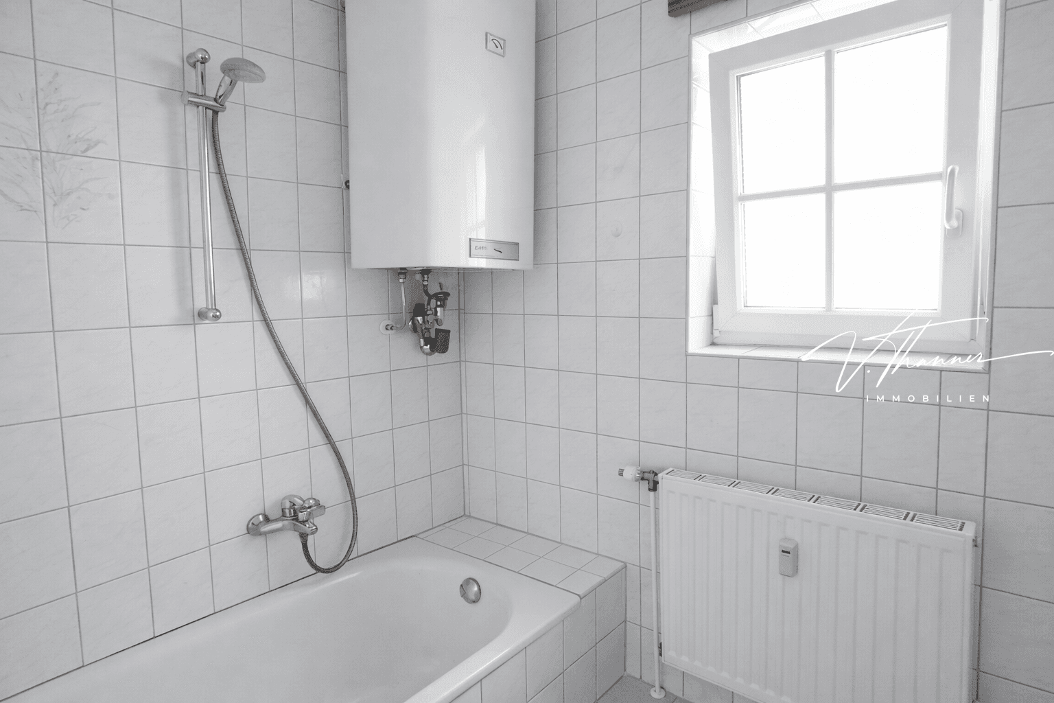 Gefliestes Badezimmer mit Badewanne, Duscharmatur, Warmwasserboiler und Fenster.