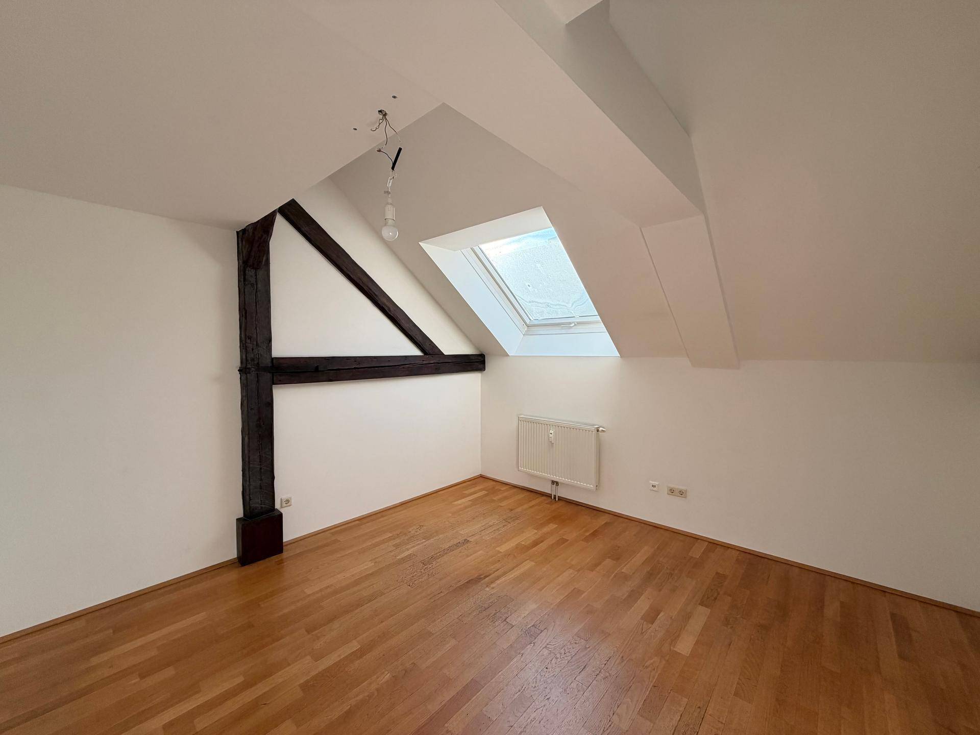 Helles Zimmer mit Holzboden, Dachschräge, Oberlicht und sichtbarem Holzbalken.