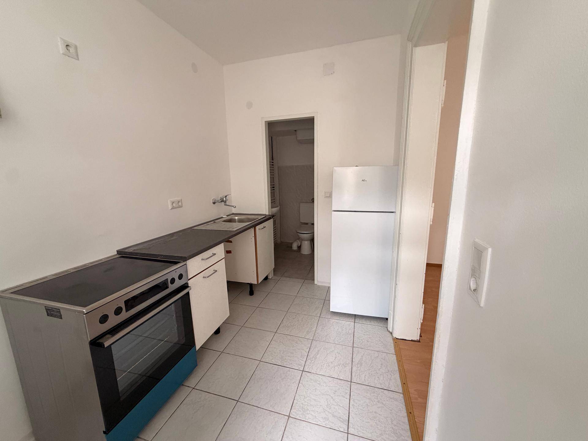 Einfache Küche mit Herd, Spüle und Kühlschrank, angrenzend an ein kleines Badezimmer.