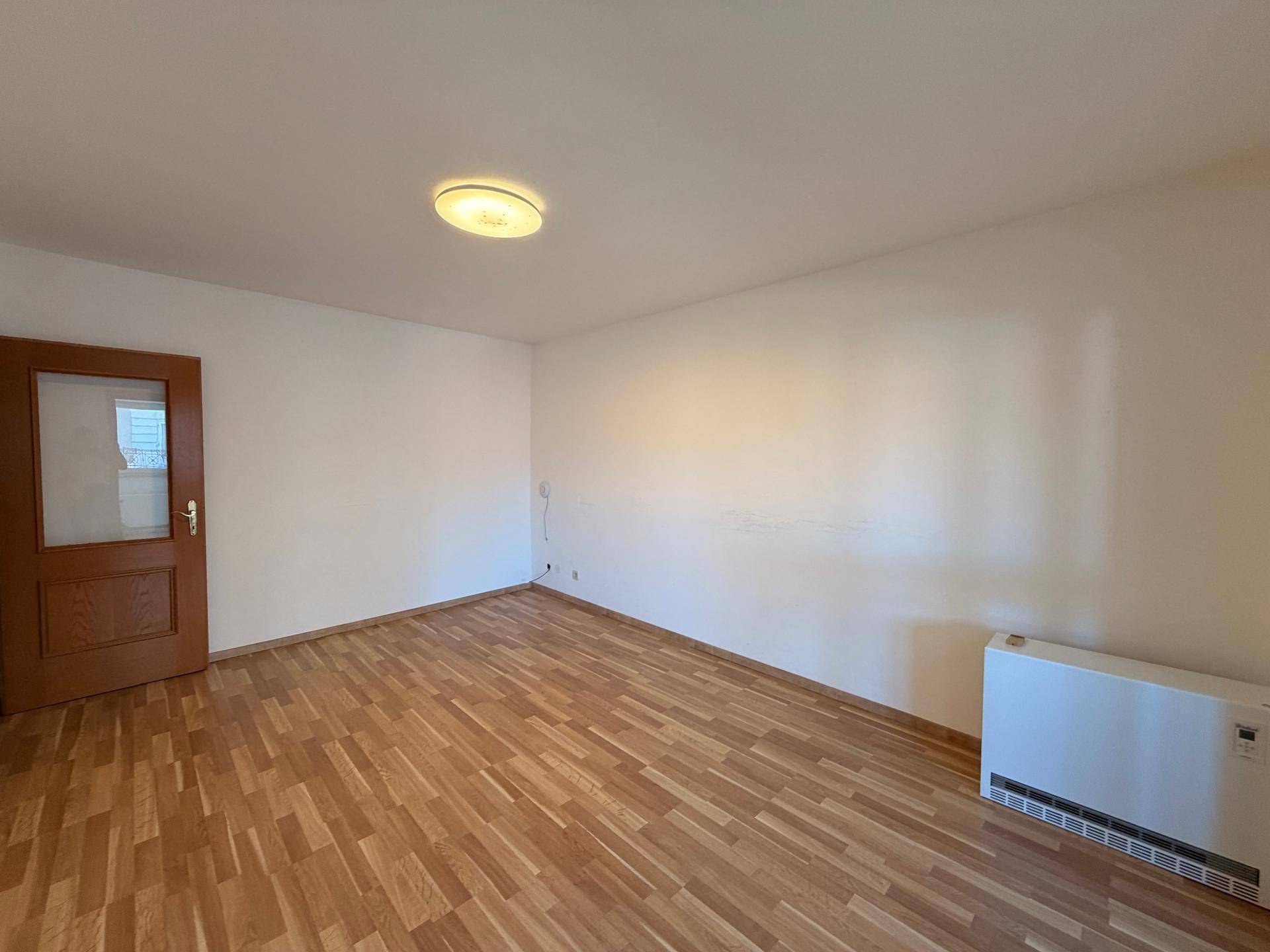 Leerer Raum mit Laminatboden, weiß gestrichenen Wänden und einem Heizkörper unter dem Fenster.