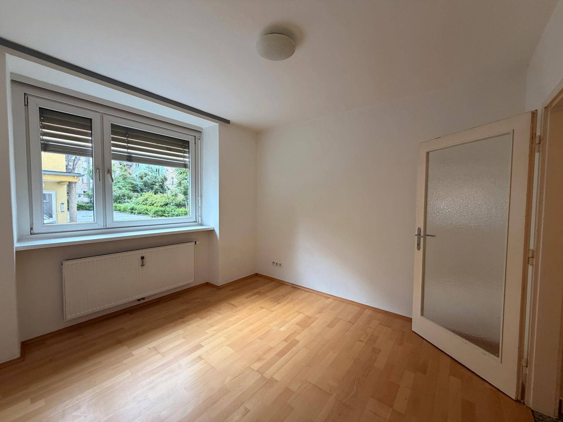 Heller Raum mit Laminatboden, einem Fenster mit Jalousien und Blick ins Grüne.