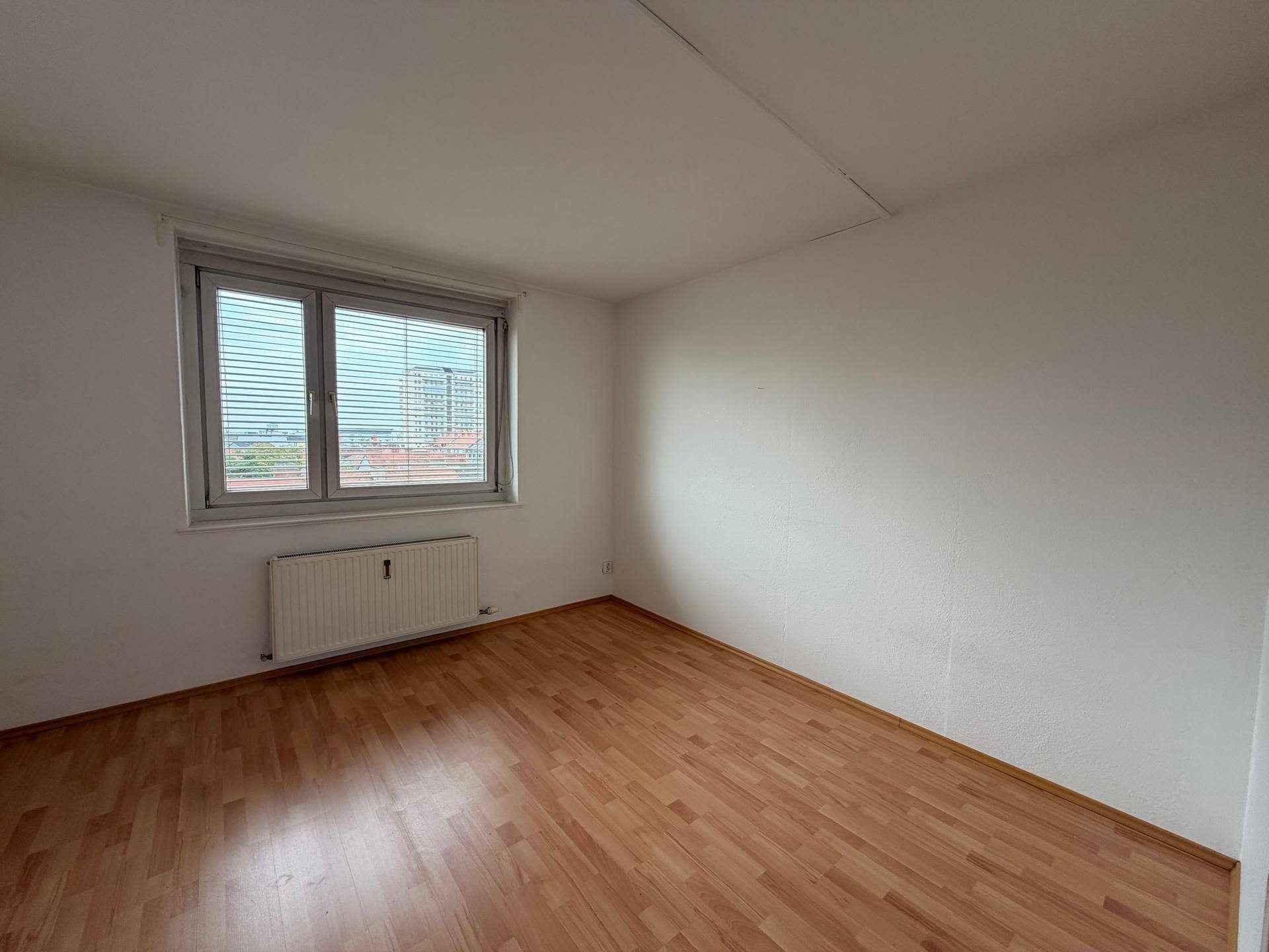Lichtdurchflutetes Zimmer mit Parkettboden und Fenster mit Jalousien und Stadtblick.