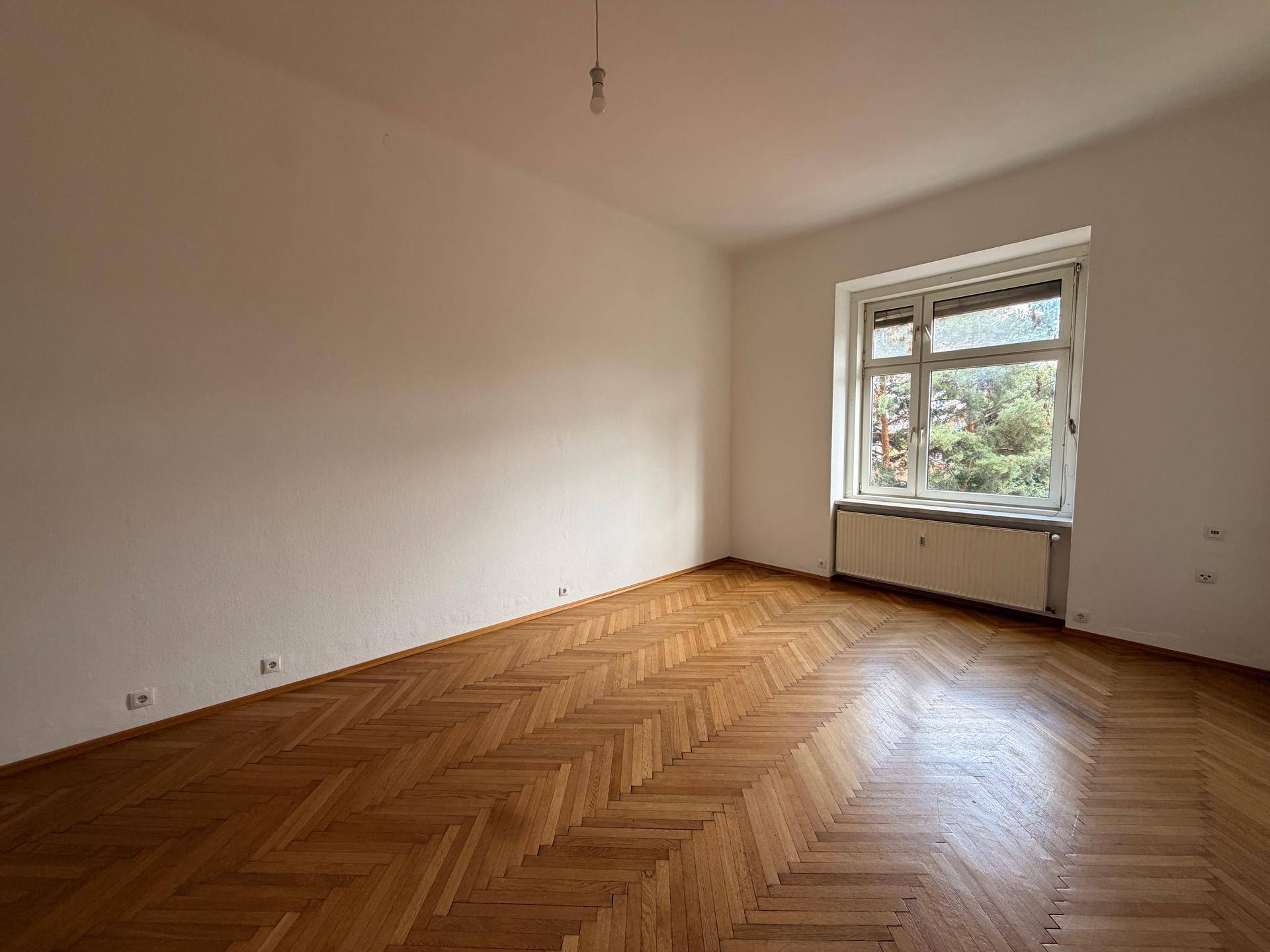 Leeres Zimmer mit hellen Wänden, einem Fenster und klassischem Fischgrätparkettboden.