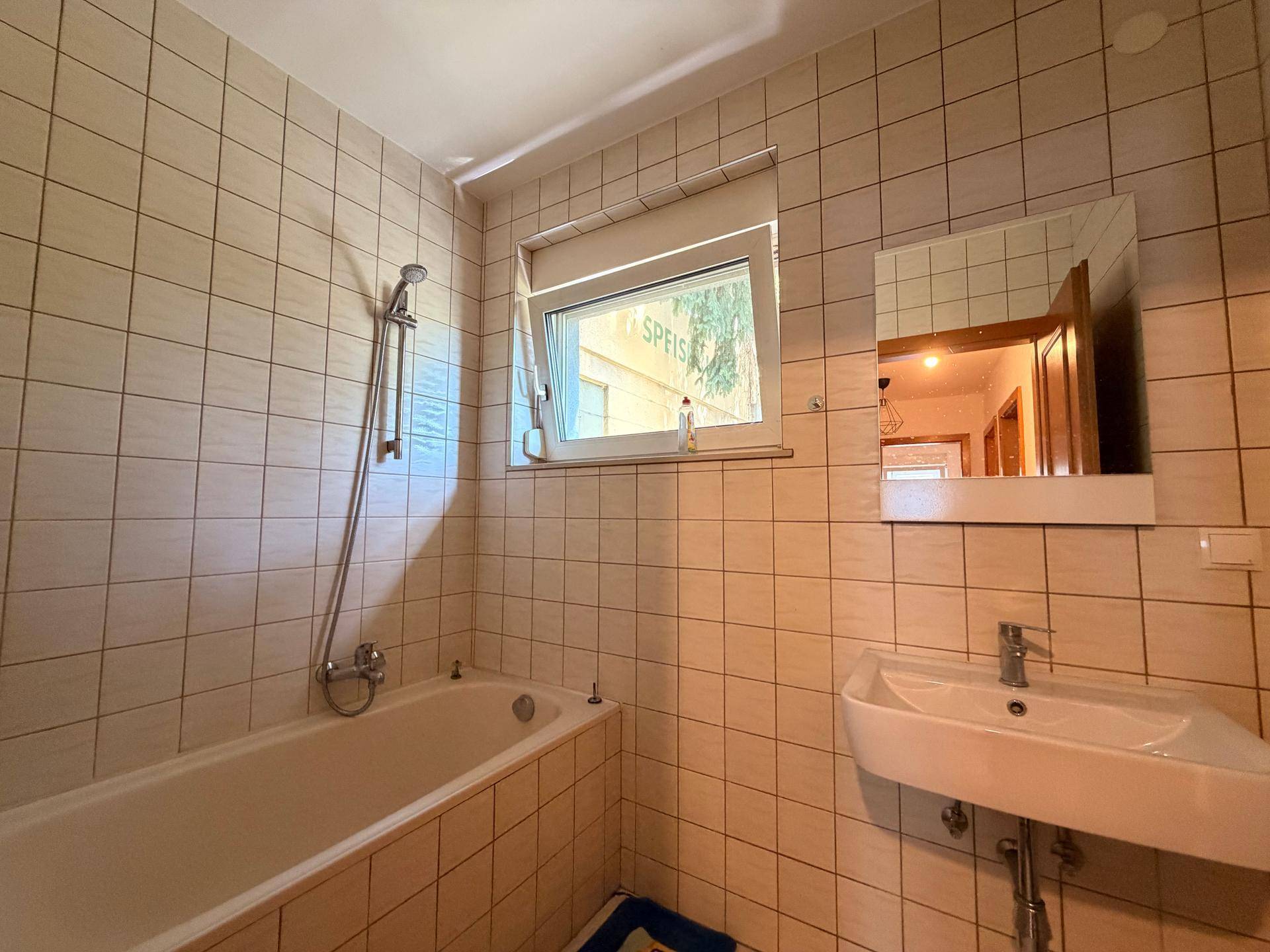 Badezimmer mit beigefarbenen Fliesen, einer Badewanne mit Duschfunktion und einem Fenster.