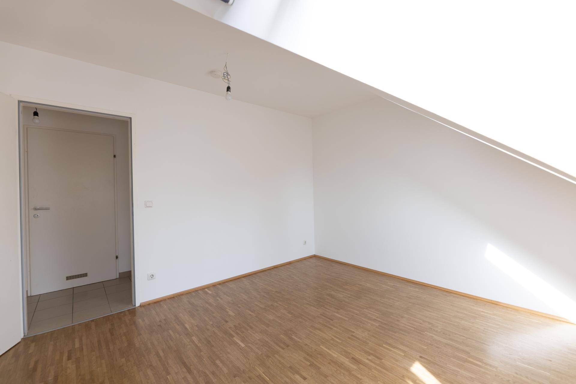 Leeres Zimmer mit Dachschräge, Parkettboden und Zugang zu einem gefliesten Bereich.