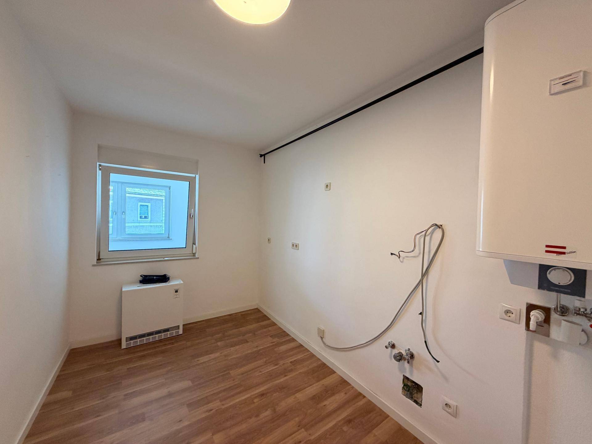 Kleiner Raum mit Laminatboden, Warmwasserbereiter und sichtbaren Installationen an der Wand.