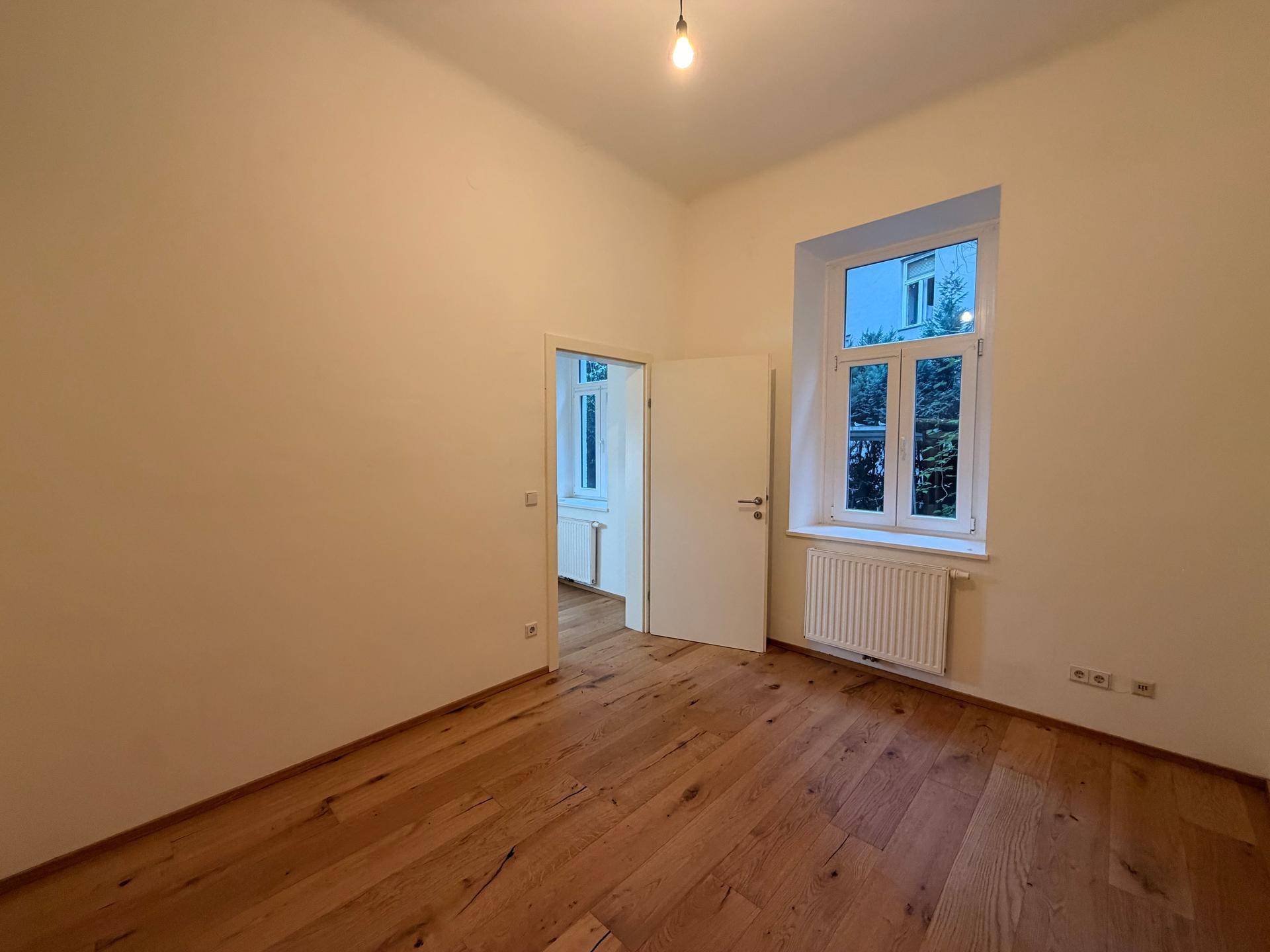 Leeres Zimmer mit Holzboden, weißen Wänden und einem großen Fenster für viel Tageslicht.