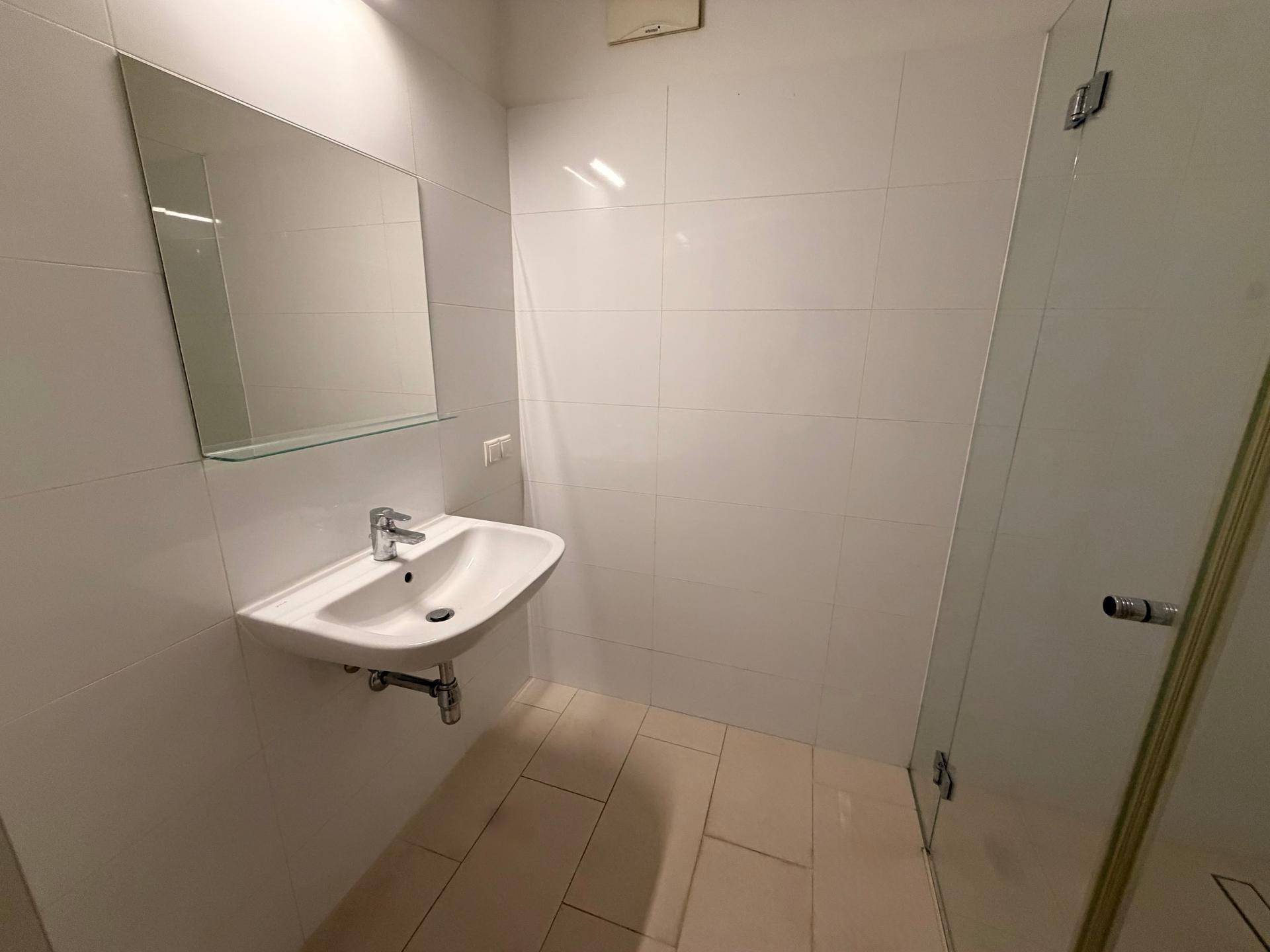 Helles Badezimmer mit Waschbecken, Spiegel und weißen Fliesen, angrenzend an eine Dusche.
