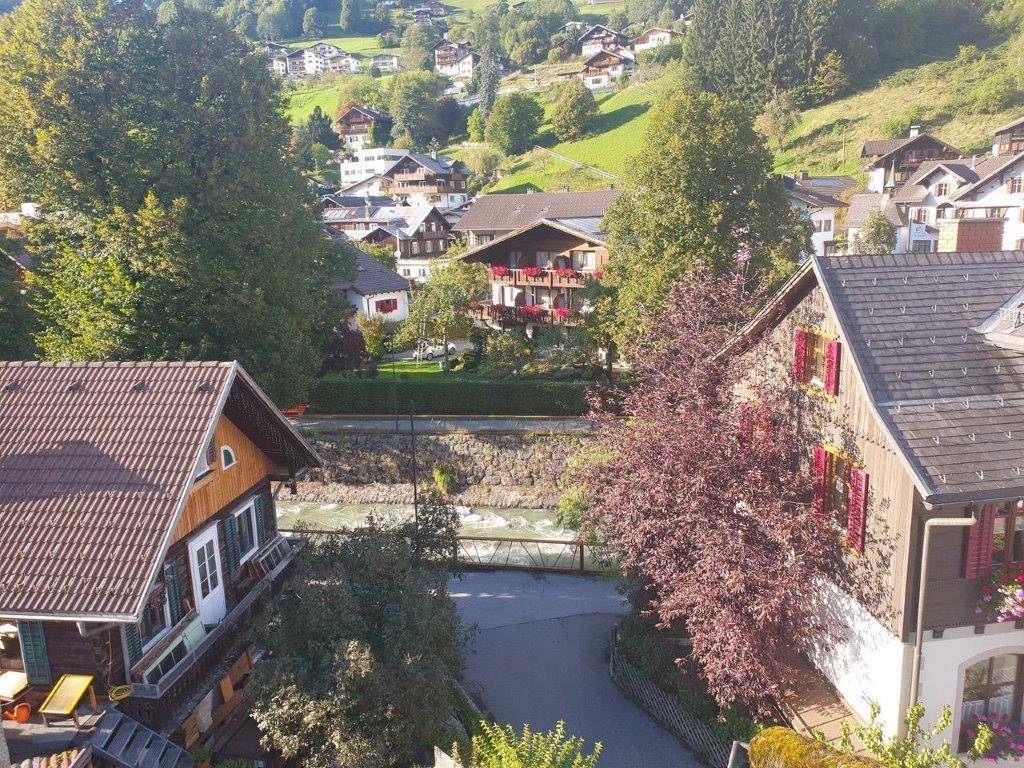 Blick auf traditionelle Häuser, einen Fluss und die grüne Hügellandschaft mit Bergen im Hintergrund.