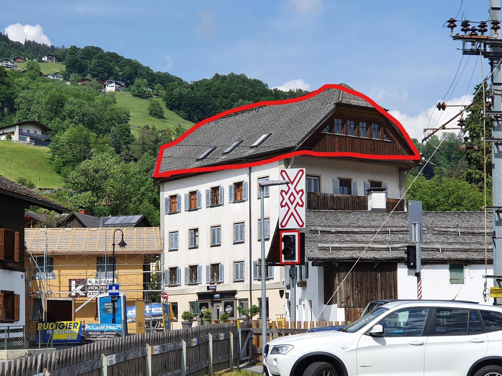 Gebäude mit markantem Mansarddach und mehreren Fenstern, umgeben von grünen Hügeln und Infrastruktur.