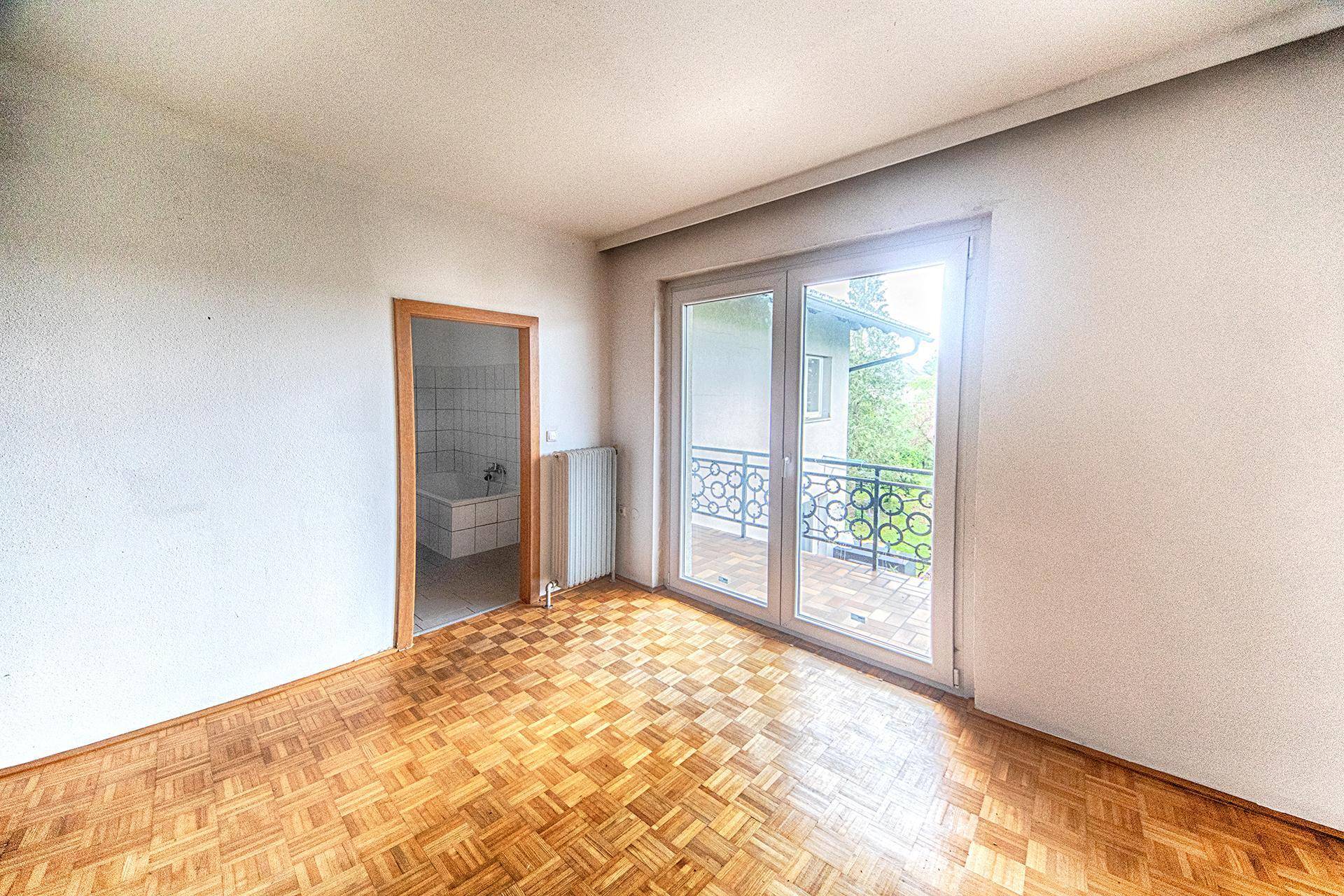 Helles Zimmer mit Parkettboden, großem Fenster und Zugang zu einem französischen Balkon sowie einem Bad.