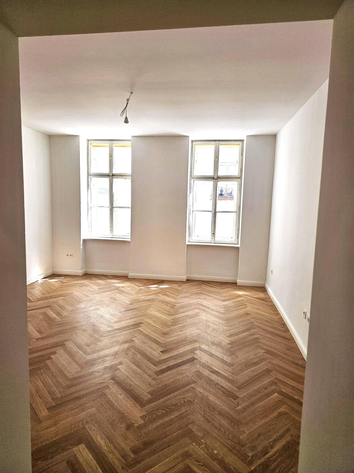 Geräumiger Raum mit klassischem Fischgrätparkett und zwei hohen Fenstern, bereit zur individuellen Einrichtung.