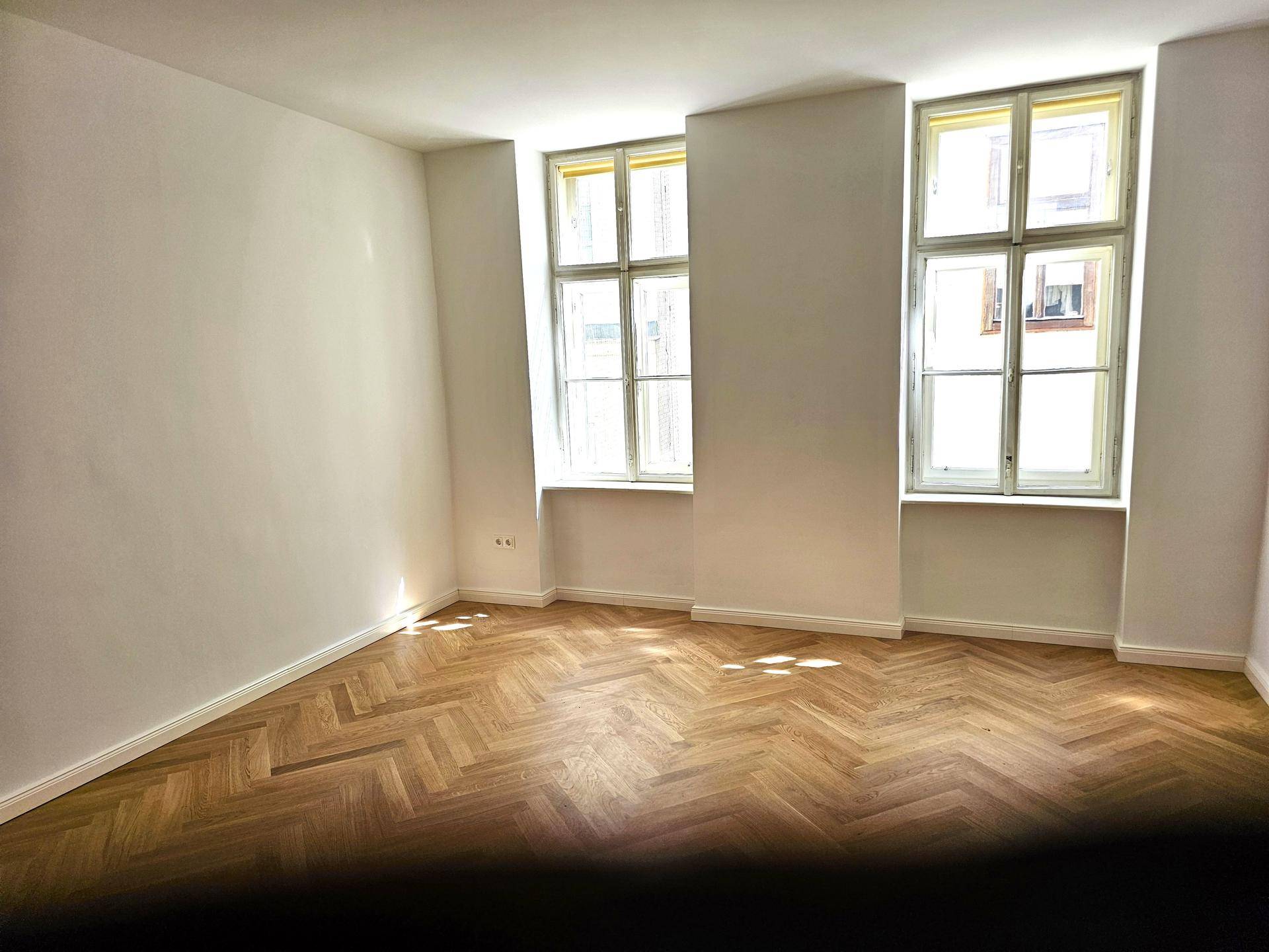 Heller Raum mit klassischem Fischgrätparkett und zwei hohen Fenstern, bietet viel Tageslicht und Raumgefühl.