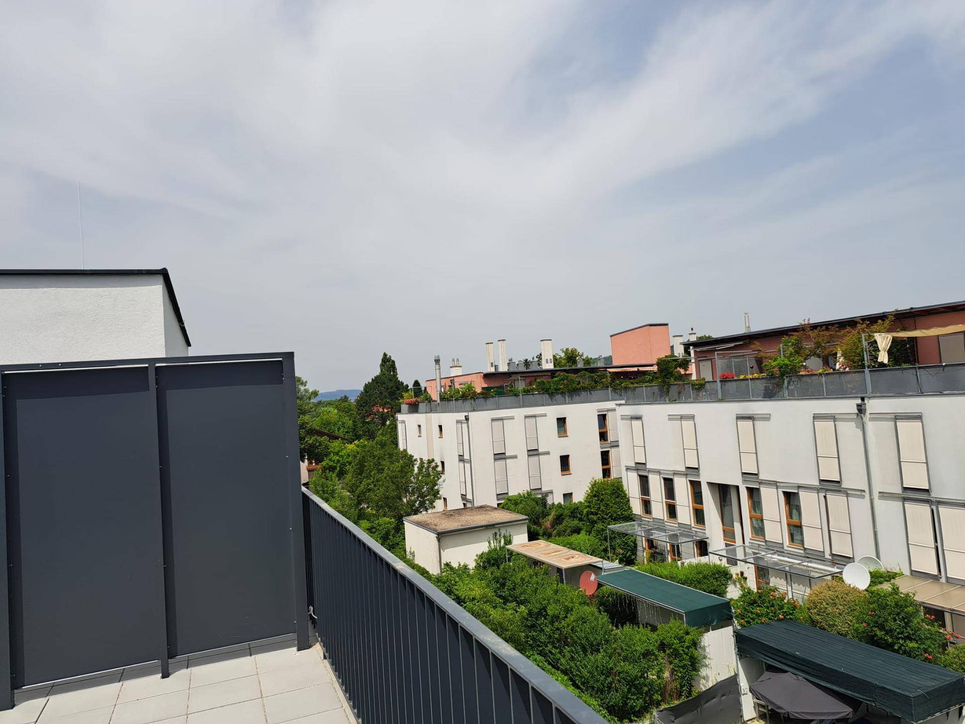 Terrasse mit Blick auf umliegende Gebäude und grüne Bäume unter einem bewölkten Himmel.