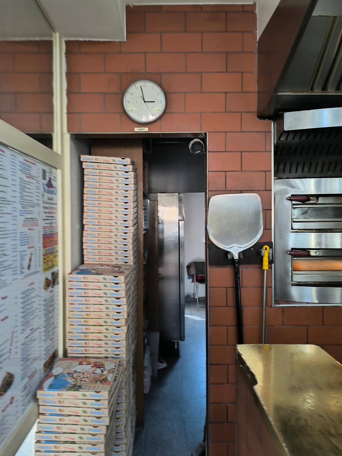 Teil einer Gewerbeküche mit gestapelten Pizzakartons, einer Uhr und einem Durchgang.
