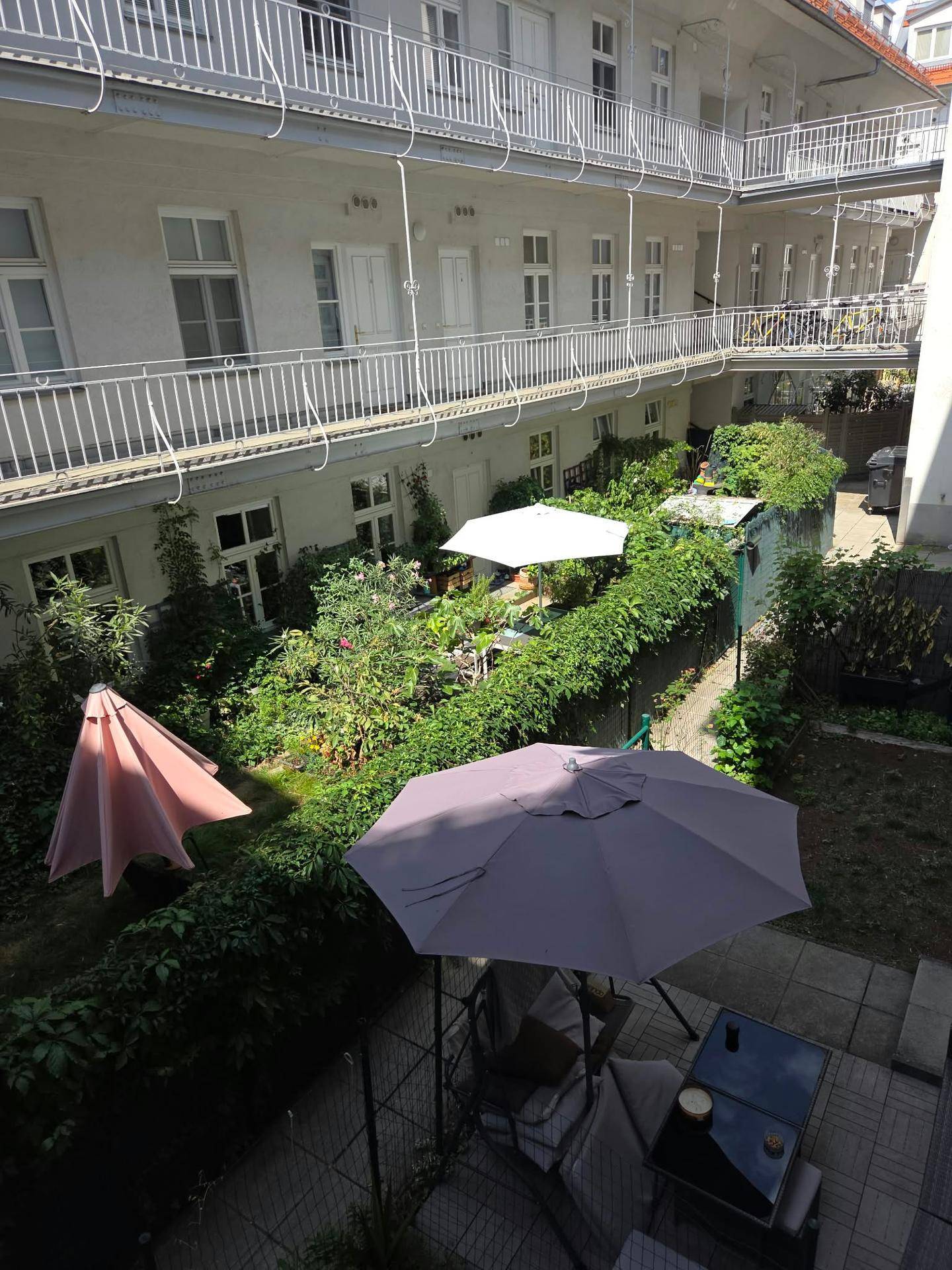 Ein grüner Innenhof mit Gartenmöbeln, Sonnenschirmen und umlaufenden Balkonen in einem Wohngebäude.
