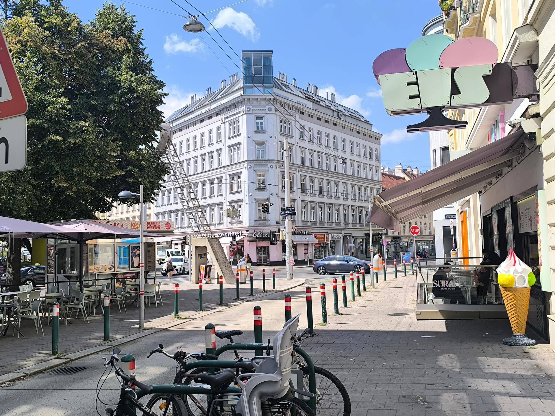Belebtes Stadtbild mit einem markanten Eckgebäude, einem Eiscafé und Fahrrädern auf dem Bürgersteig.