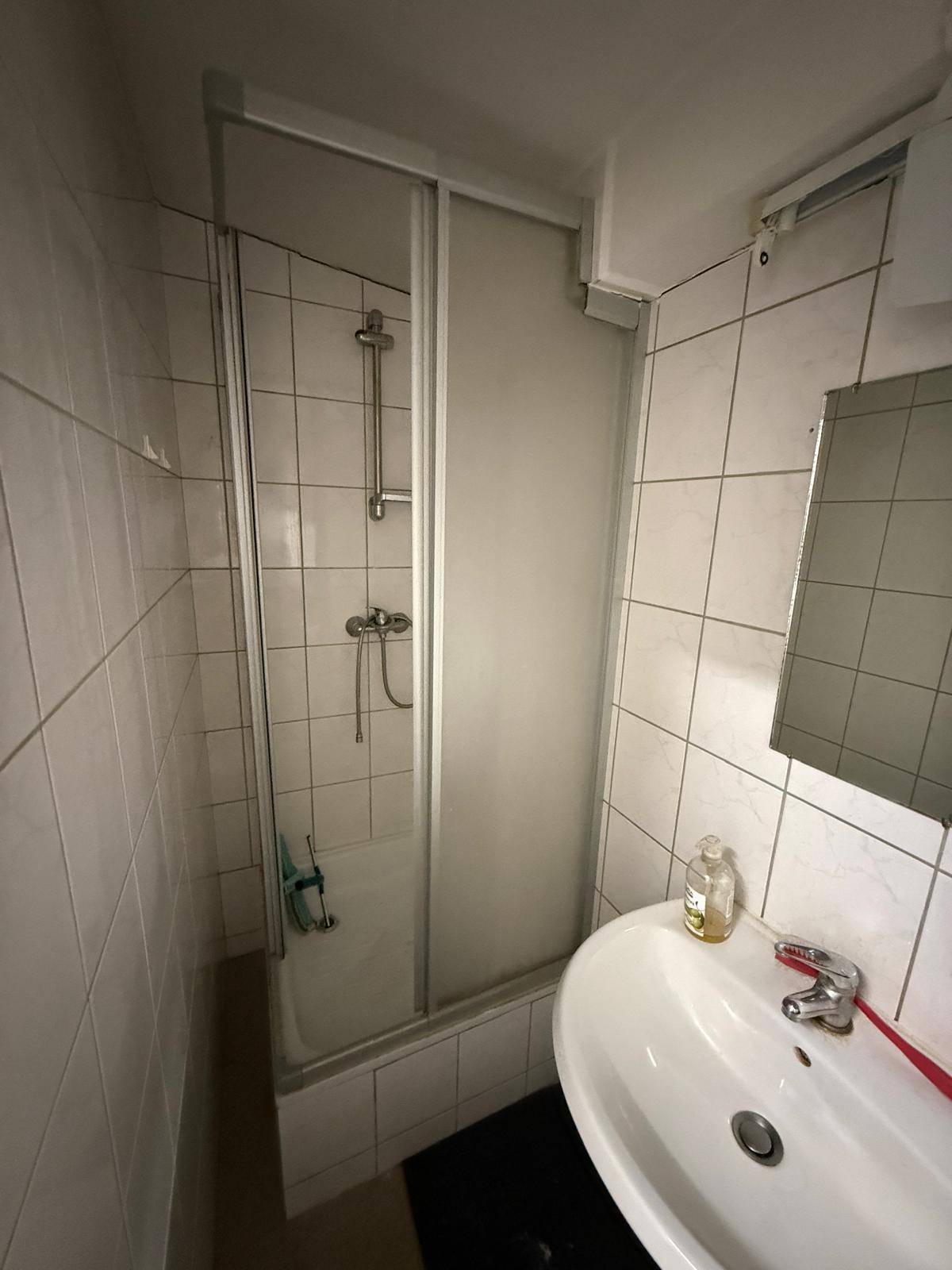 Kleines Badezimmer mit Duschkabine, Waschbecken und weißen Fliesen an den Wänden.