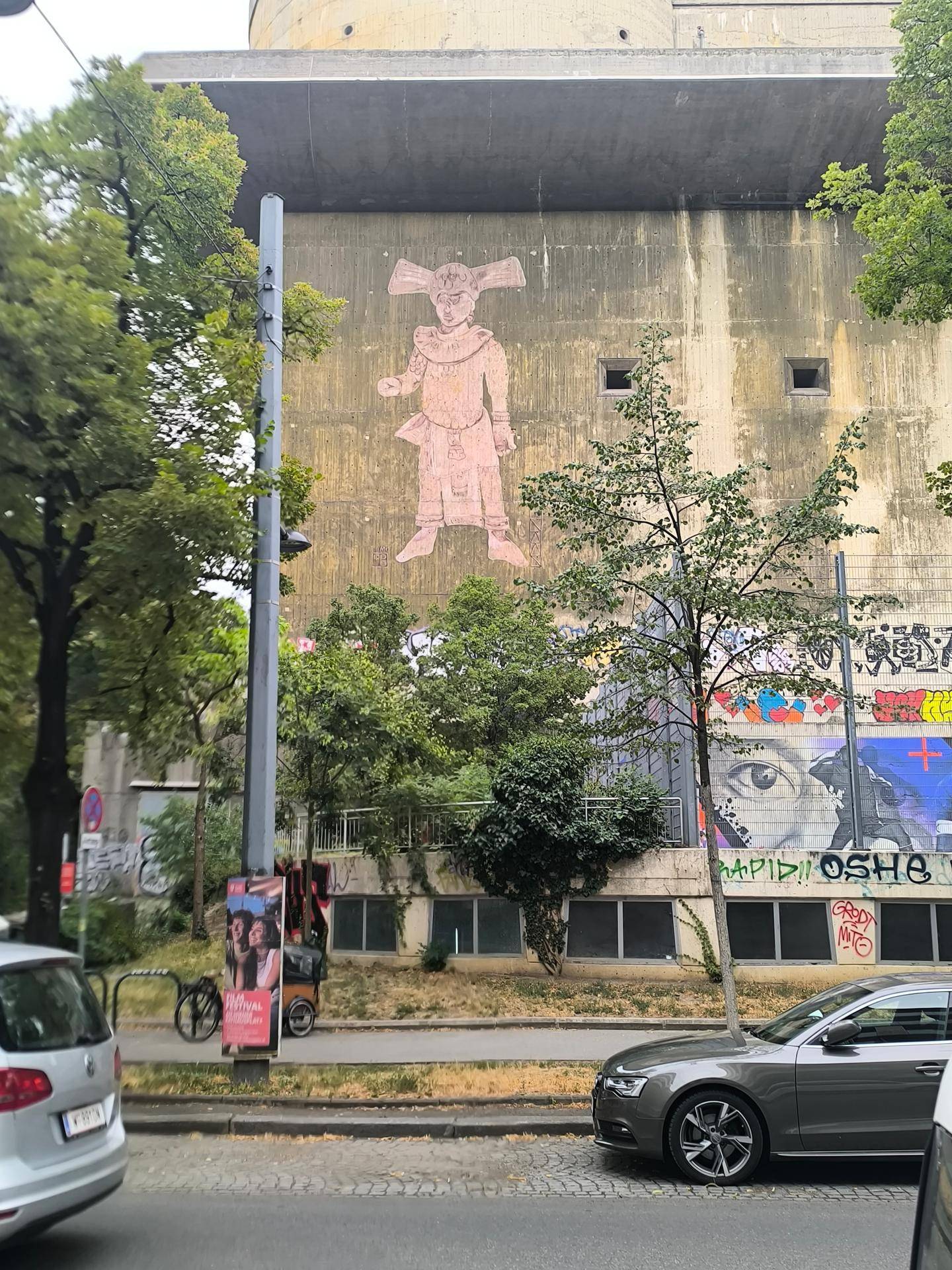 Massives Betongebäude mit Graffiti und Bäumen an einer Straße, mit parkenden Autos im Vordergrund.