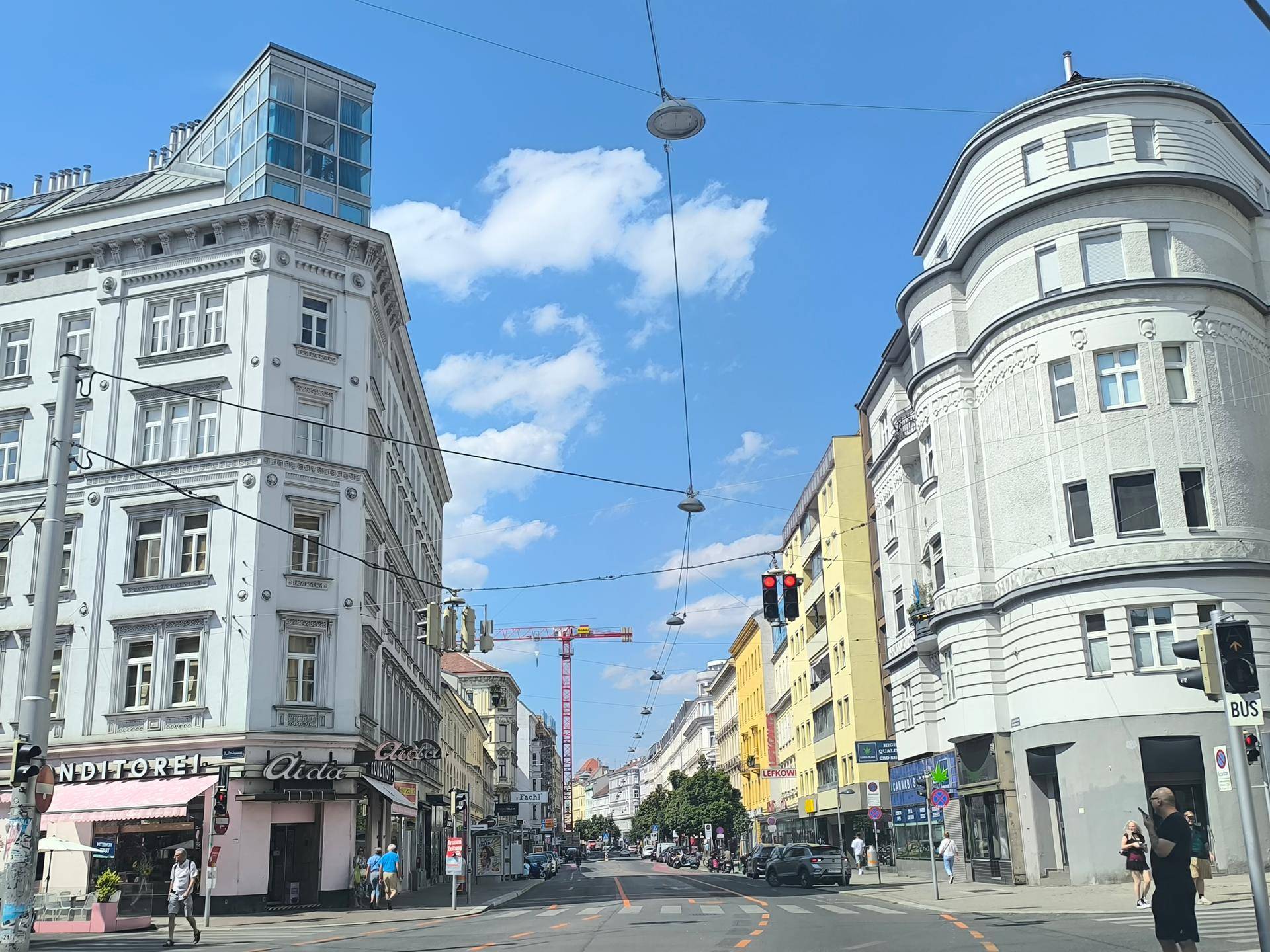 Weitläufige Straßenansicht einer Stadt mit historischen Gebäuden, Verkehr und Fußgängern unter blauem Himmel.