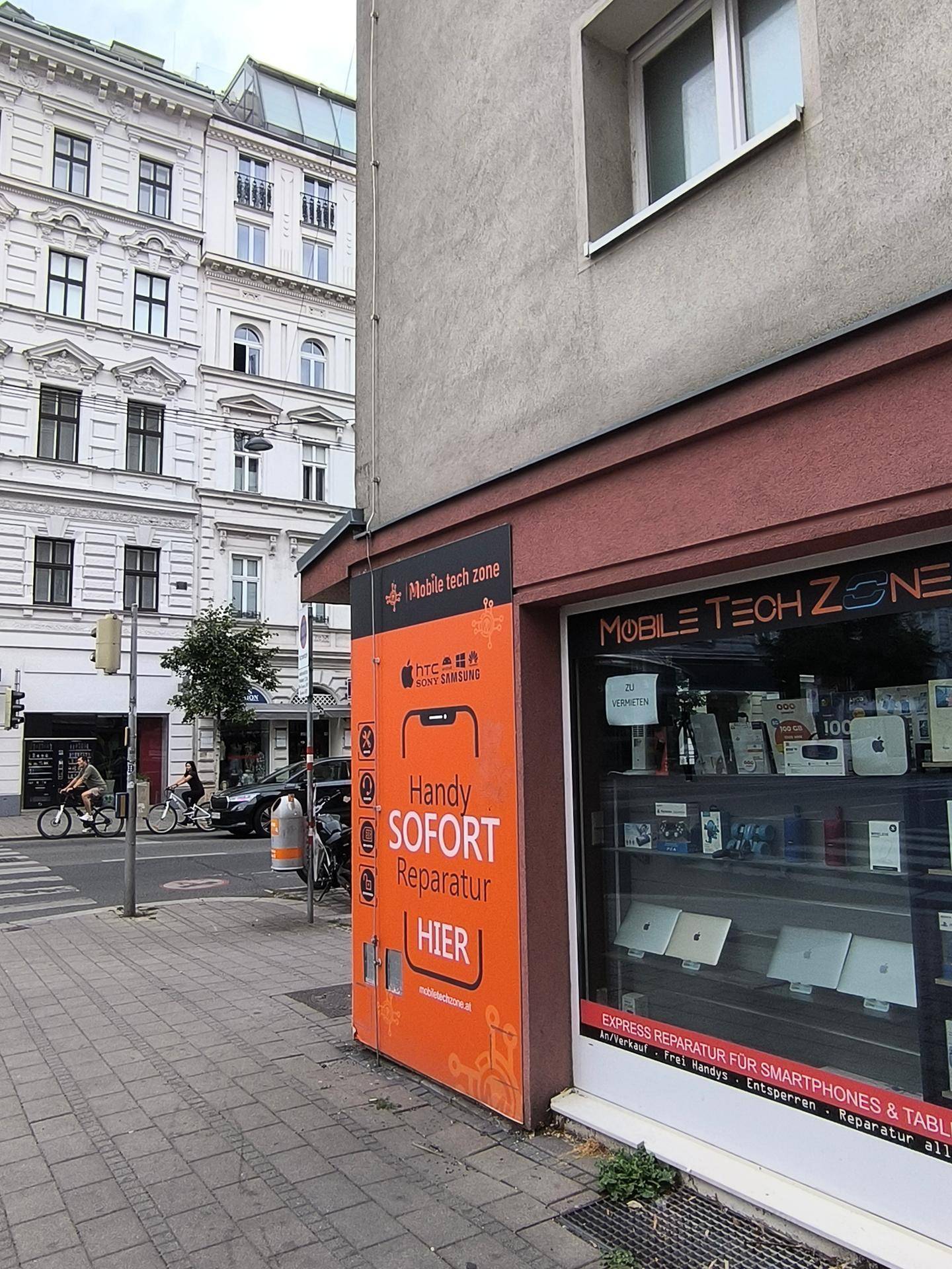 Seitenansicht eines Technikgeschäfts an einer Straße, mit einem historischen Gebäude im Hintergrund.