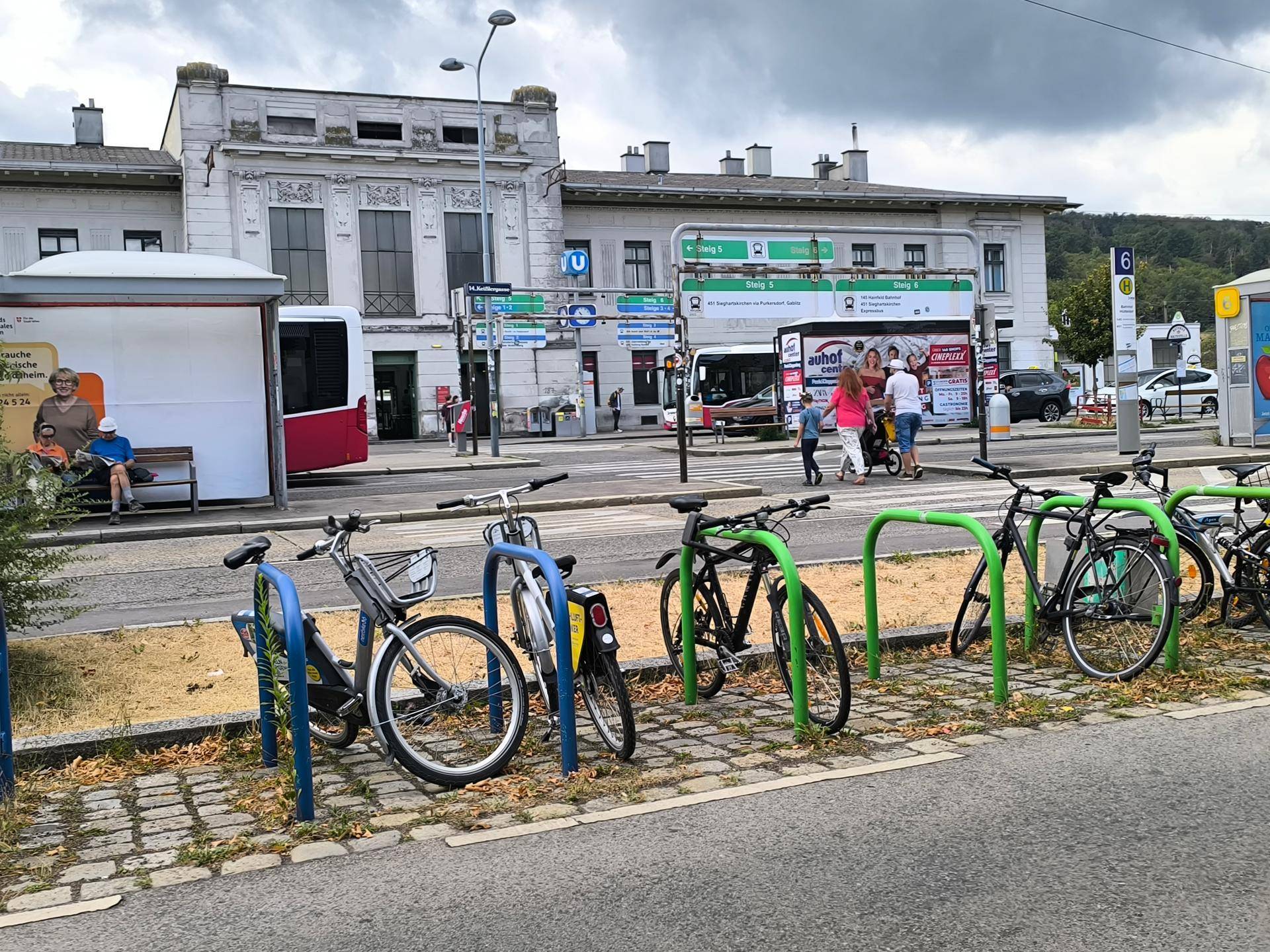 Eine Straßenszene mit einem historischen Gebäude, Bussen und Fahrradständern im Vordergrund, belebt von Passanten.