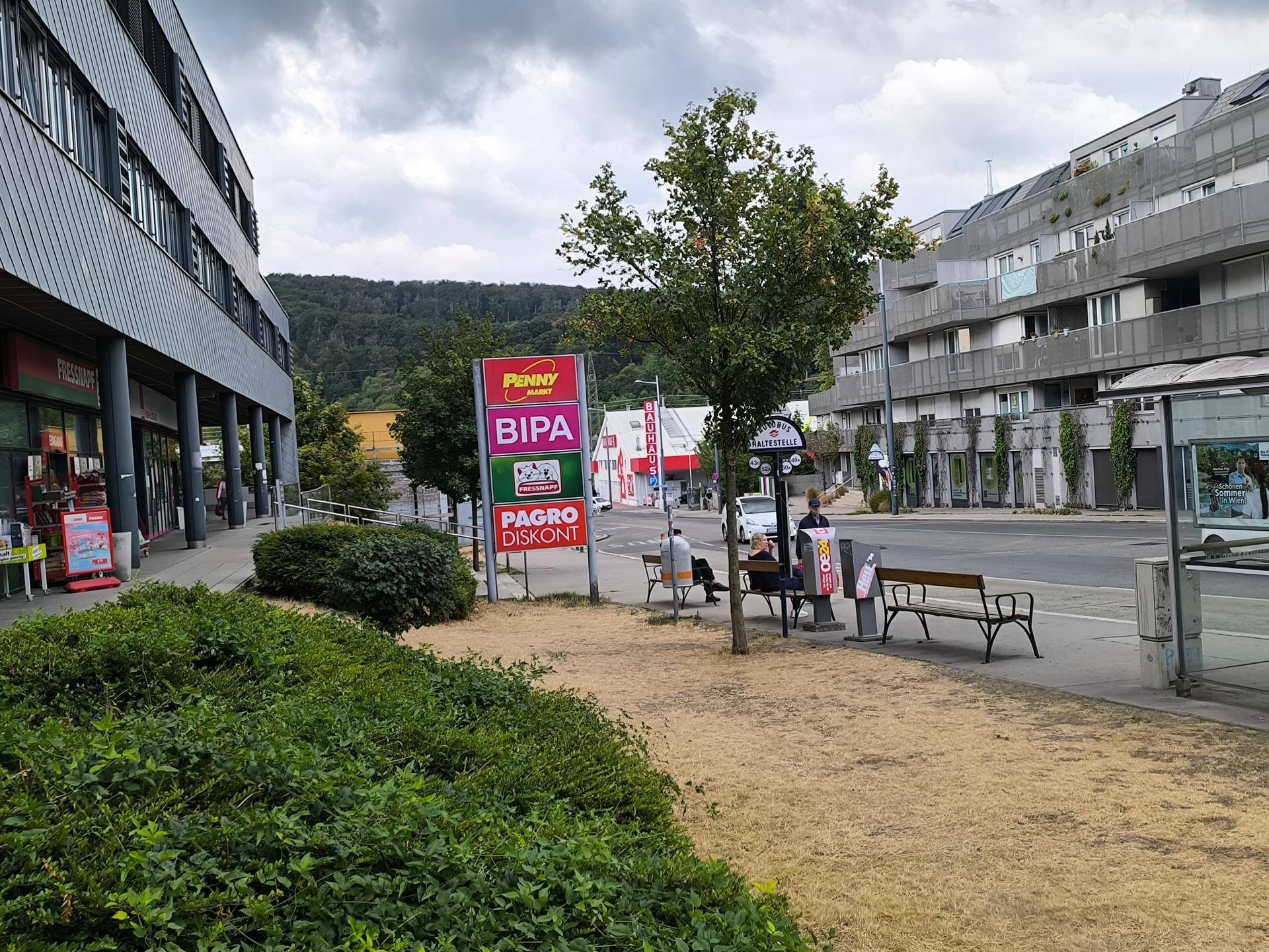Eine belebte Straßenszene mit Geschäften, Bäumen und Bänken, im Hintergrund sind grüne Hügel zu sehen.