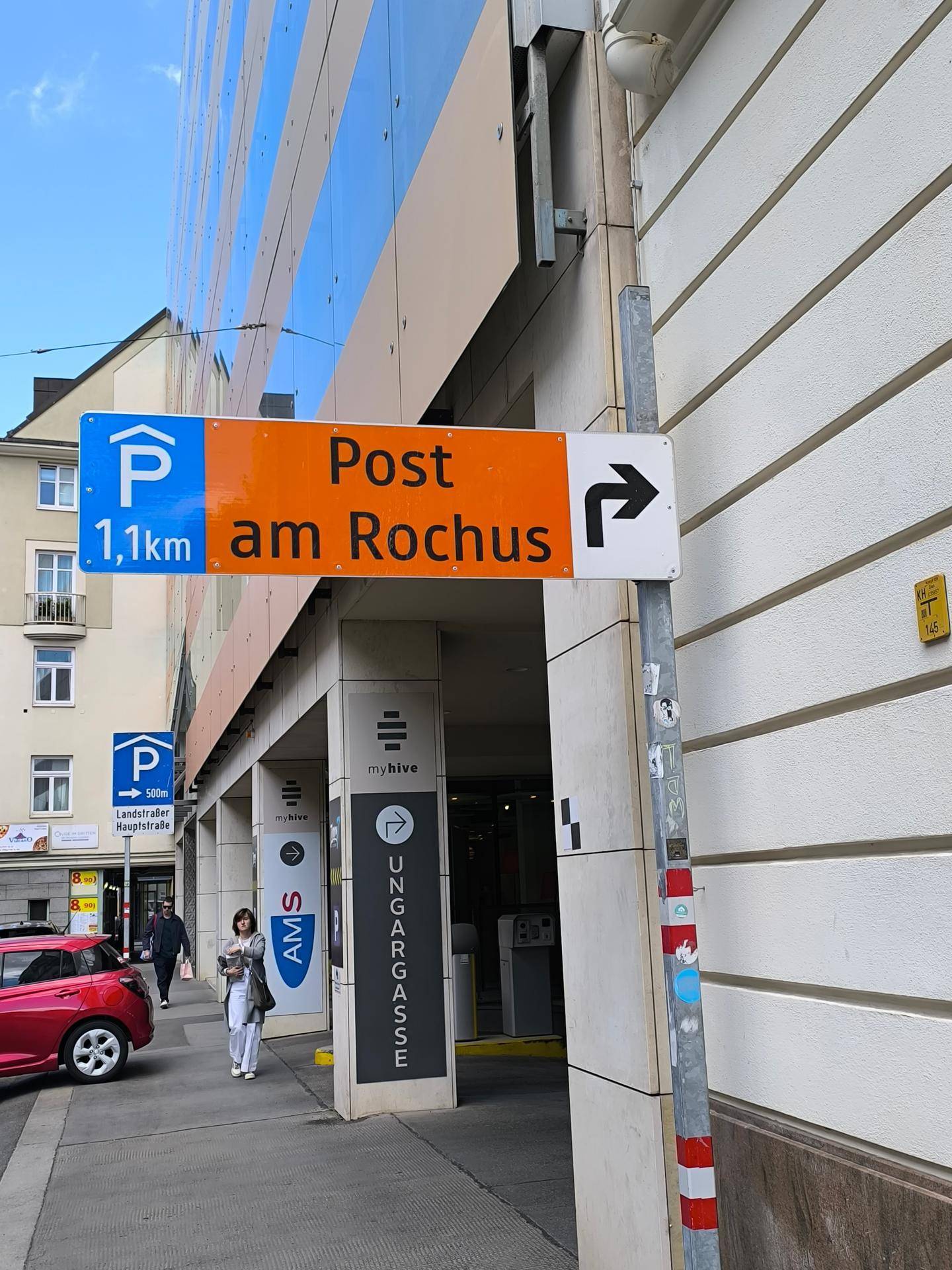 Straßenschilder weisen auf eine Tiefgarage und die Post am Rochus hin, vor einer modernen Gebäudefassade.