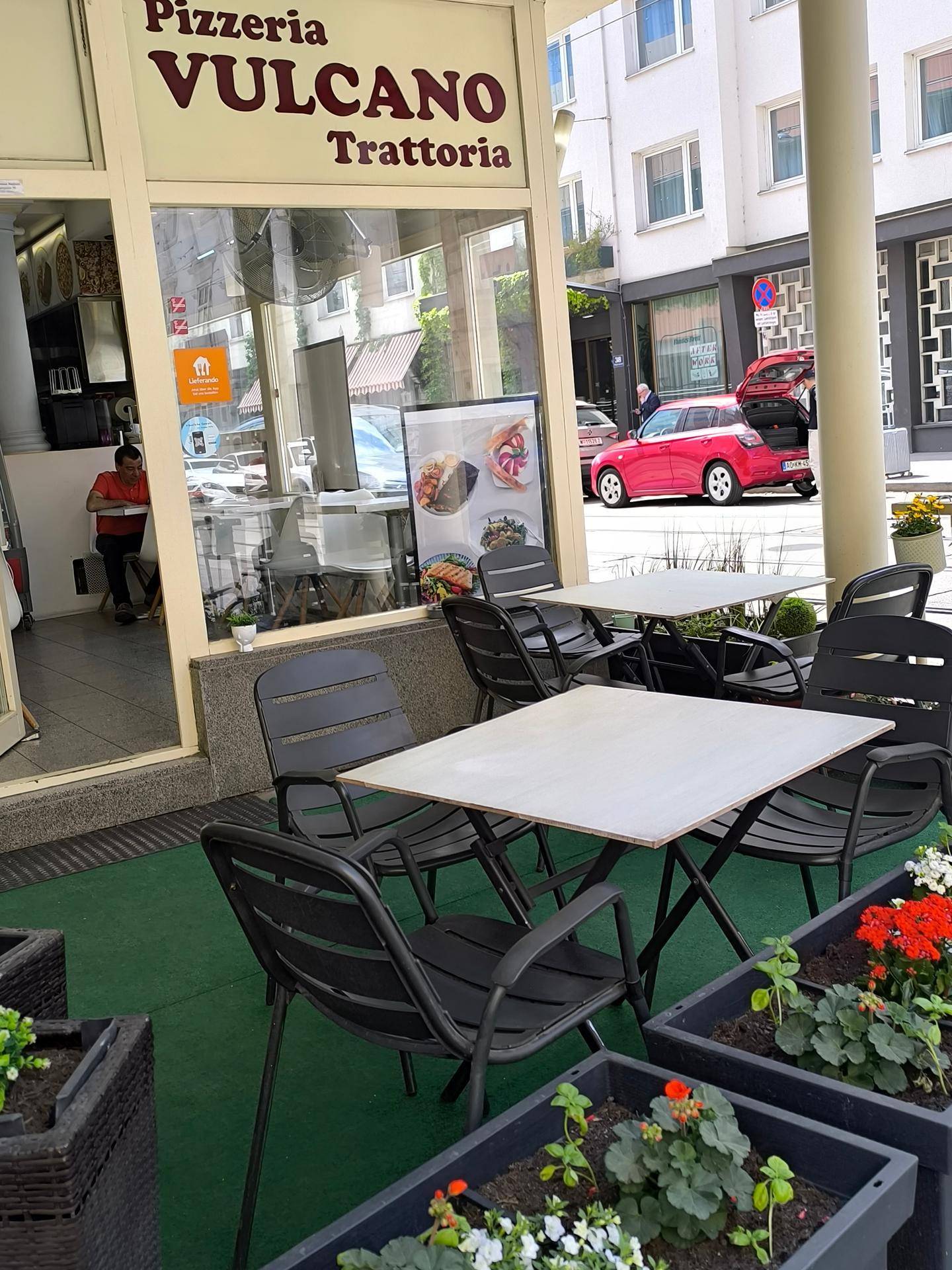 Außenterrasse eines Restaurants mit Tischen und Stühlen, umgeben von Blumenkästen in einer städtischen Umgebung.