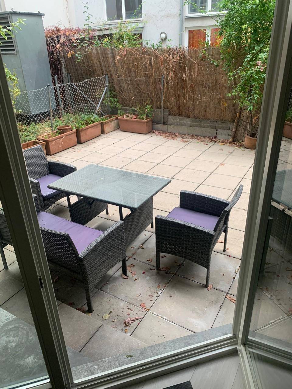 Gemütliche Terrasse mit Rattanmöbeln und Blick auf einen begrünten Hinterhofbereich mit Hecke.