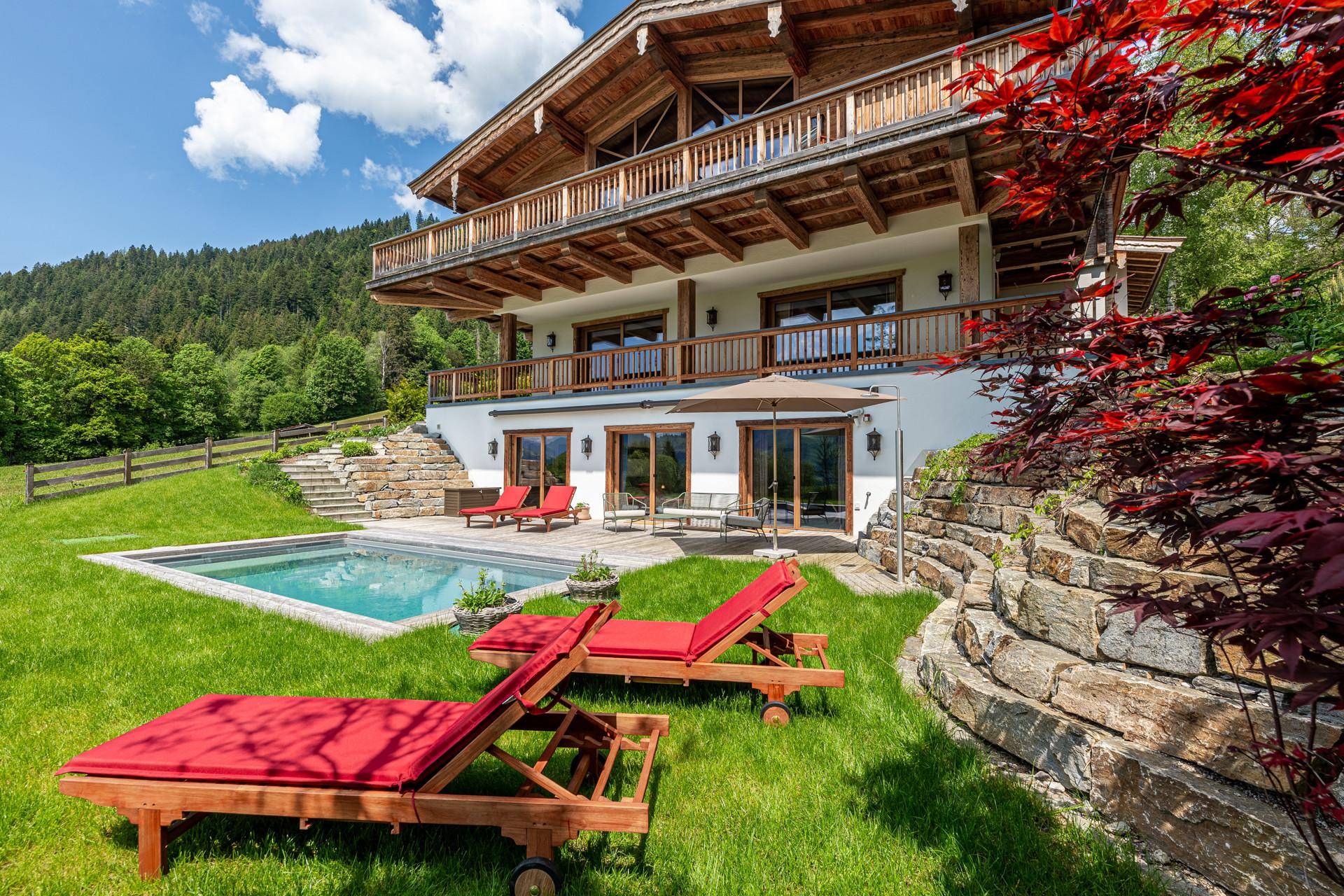Sonniger Gartenbereich mit Pool, Liegestühlen und traditioneller Steinmauer vor bewaldeter Kulisse.