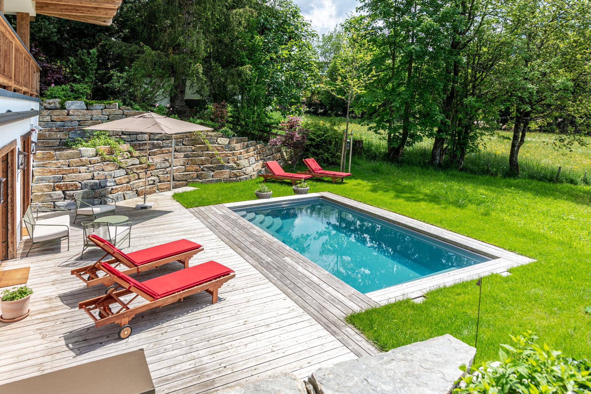 Außenbereich mit rechteckigem Pool, Holzdeck, roten Liegestühlen und einer Natursteinmauer im Grünen.