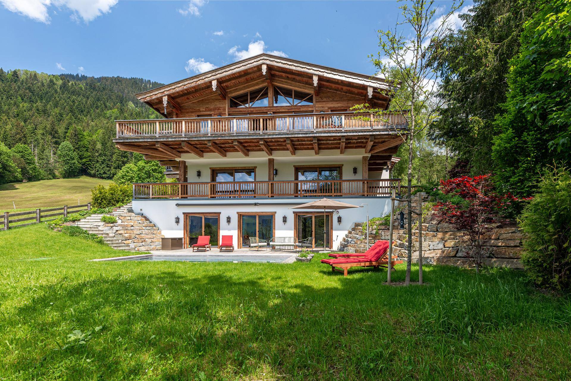 Gesamtaufnahme des Chalet-Hauses mit mehreren Balkonen, umgeben von grünen Wiesen und bewaldeten Hügeln.