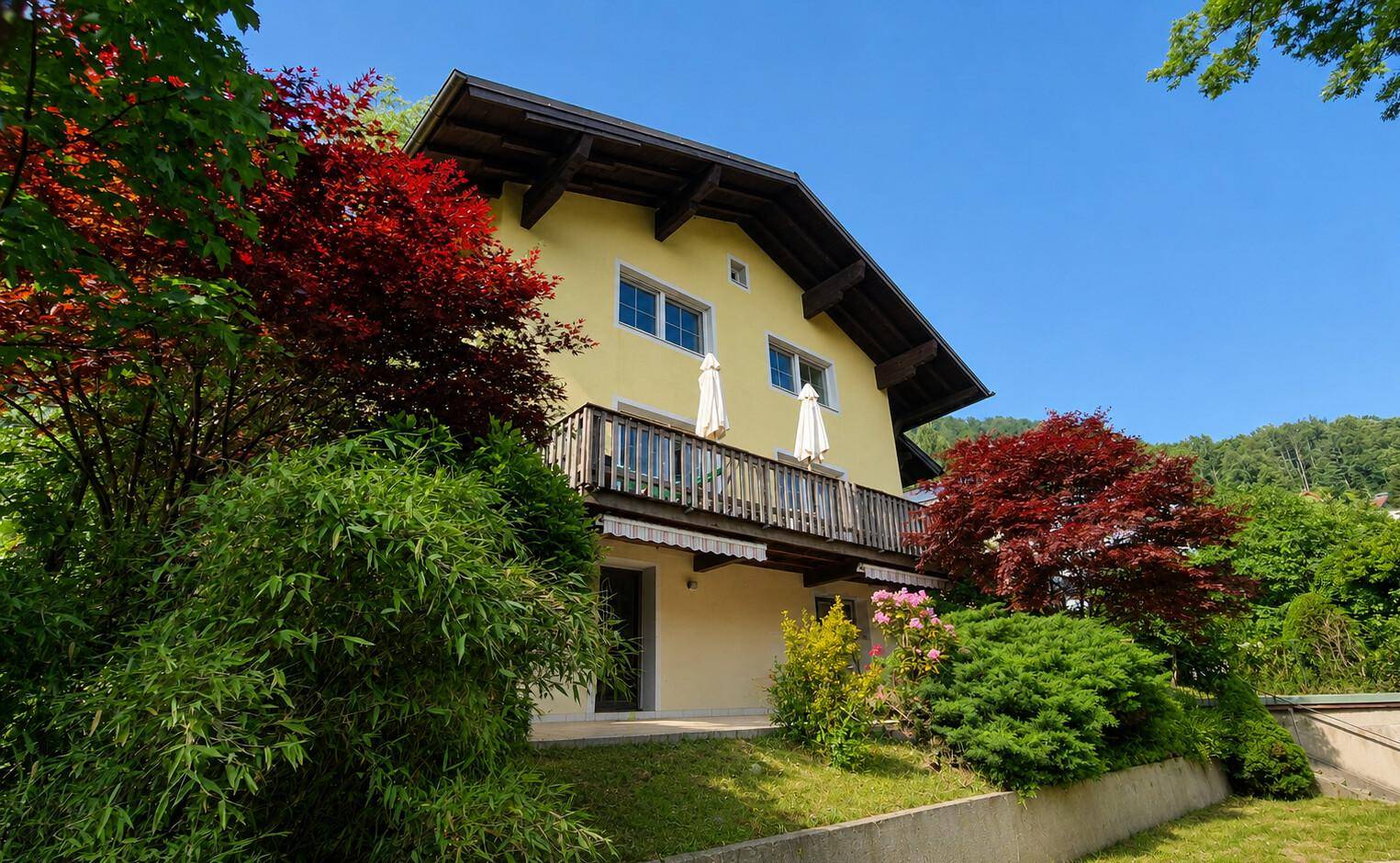 Gelbes Haus mit Balkon, ausgestattet mit zwei weißen Sonnenschirmen und umgeben von üppigem Grün.