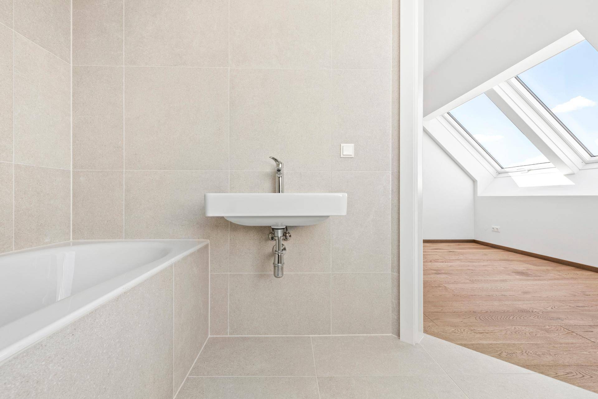 Helles Badezimmer mit Badewanne, kleinem Waschbecken und Zugang zu einem Raum mit Dachfenstern.