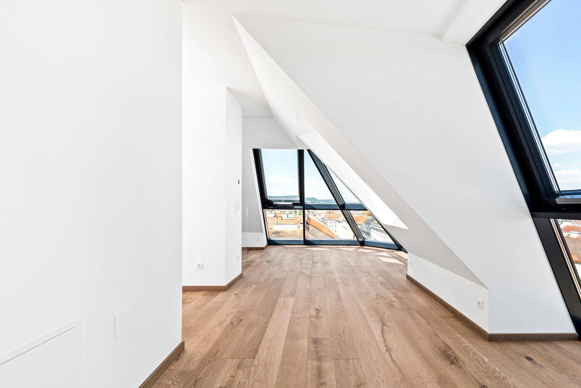 Heller, leerer Raum im Dachgeschoss mit Holzboden und großen, schrägen Fenstern mit Stadtblick.