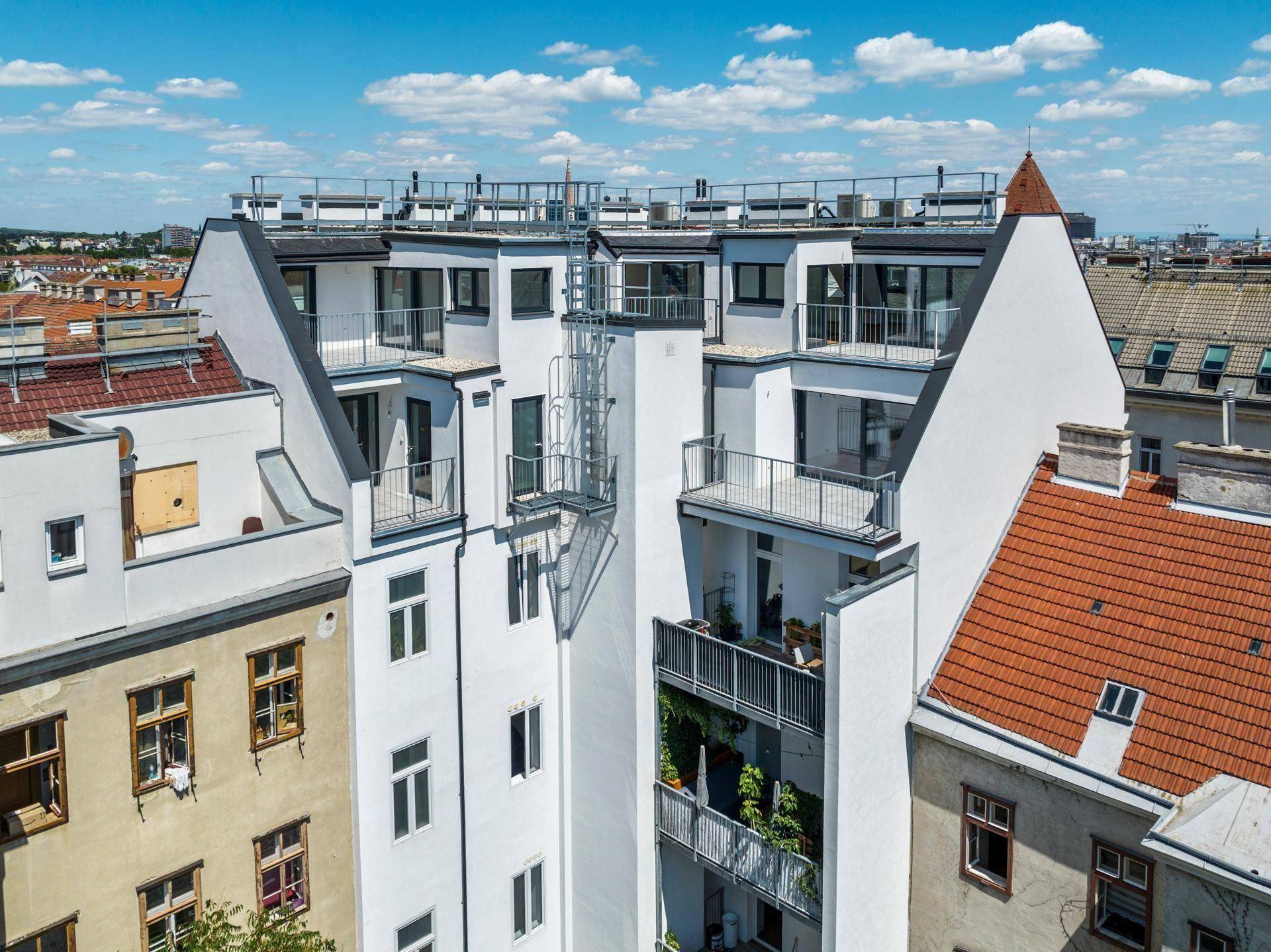 Rückansicht eines modernen Gebäudes mit mehreren Balkonen und Terrassen, umgeben von älteren Bauten.