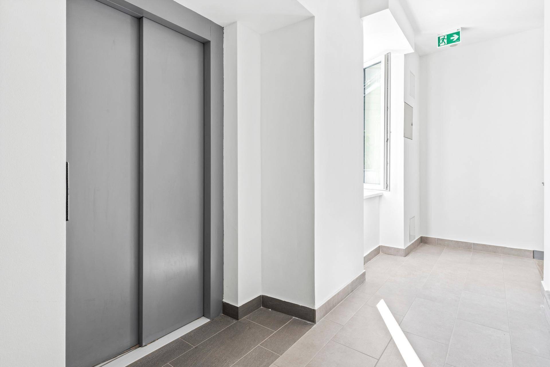 Heller Flur mit Aufzug, Fliesenboden und einem Fenster, das Tageslicht hereinlässt.