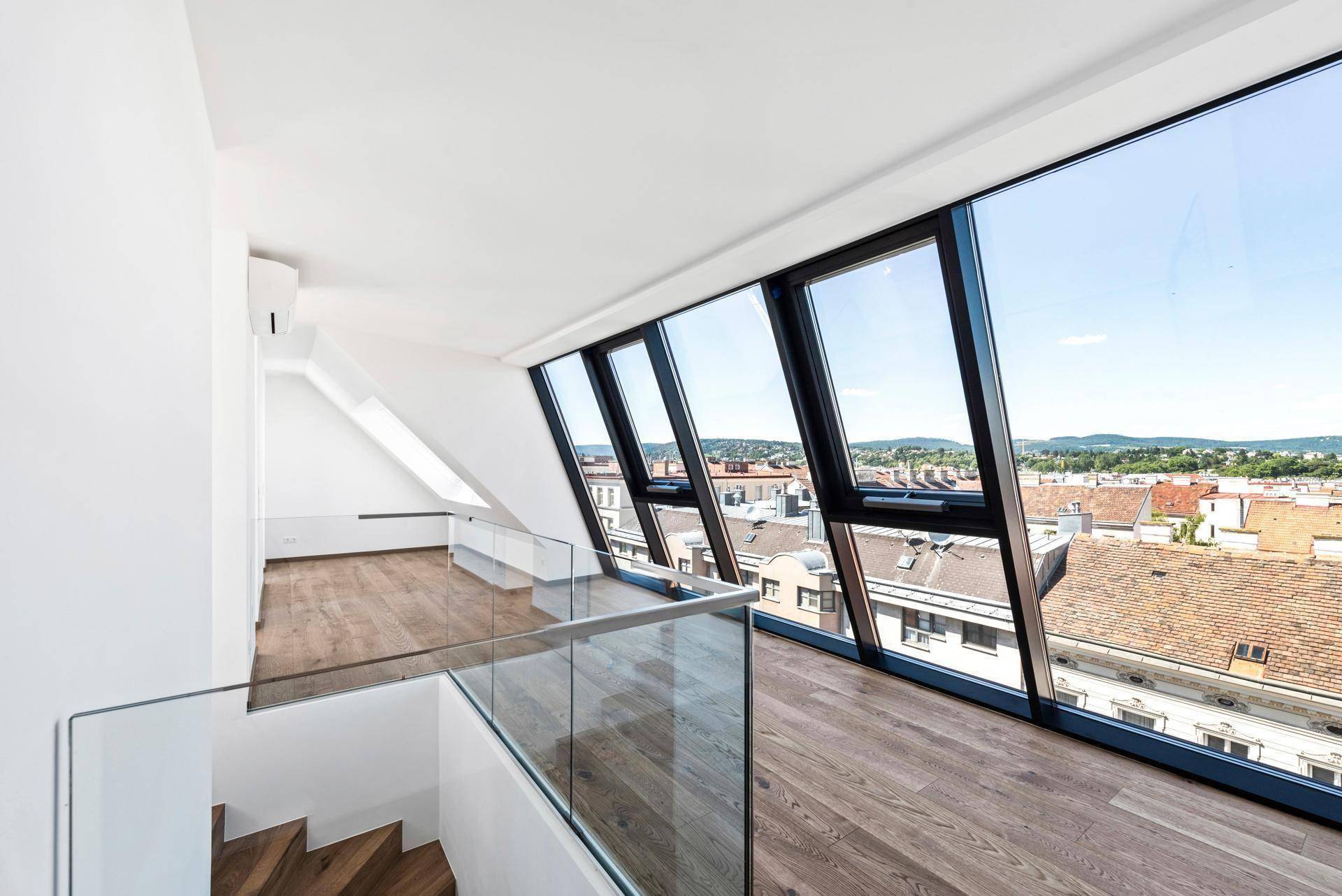 Offener Dachgeschossbereich mit Holzboden, großen Fenstern und Blick auf die Stadt, verbunden durch eine Treppe.