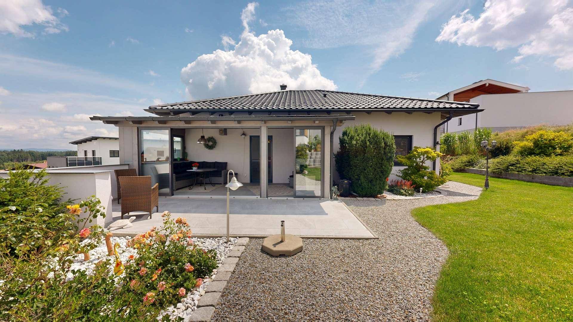 Blick vom Garten auf das Haus mit großer Terrasse, blühenden Beeten und weitläufiger Rasenfläche.