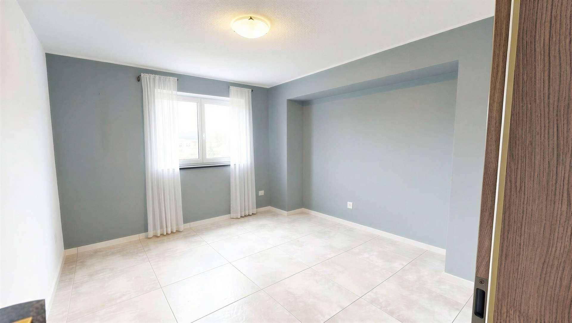 Leeres Schlafzimmer mit grauen Wänden, Fliesenboden und einem Fenster mit hellen Vorhängen.