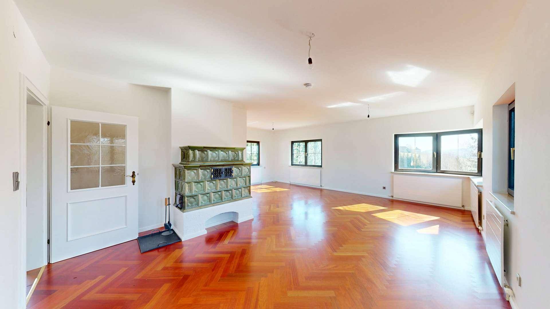 Helles Wohnzimmer mit Fischgrätparkett, traditionellem Kachelofen und großen Fenstern.