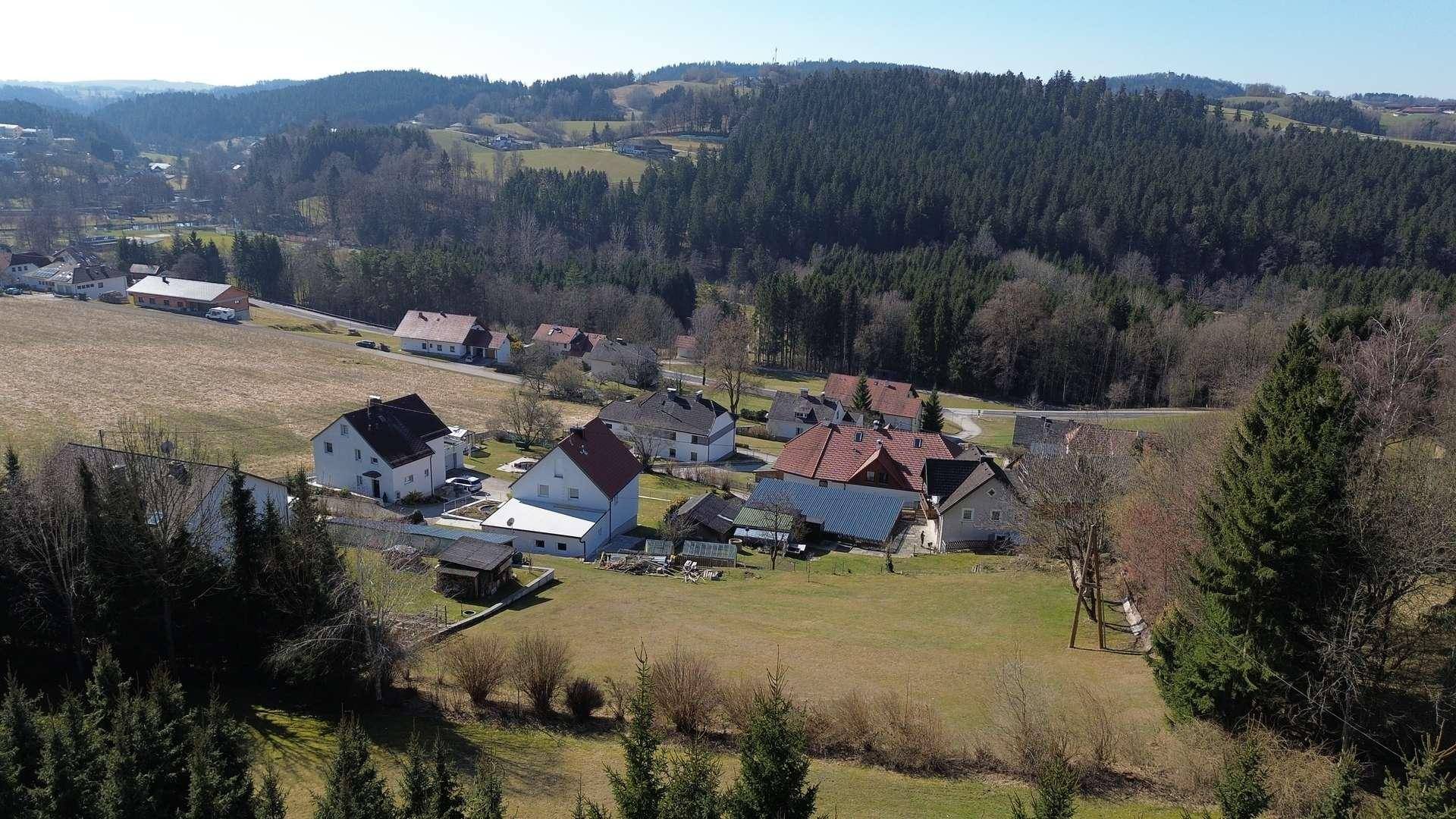 Panoramablick auf eine ländliche Siedlung in einem Tal, umgeben von bewaldeten Hügeln und Feldern.