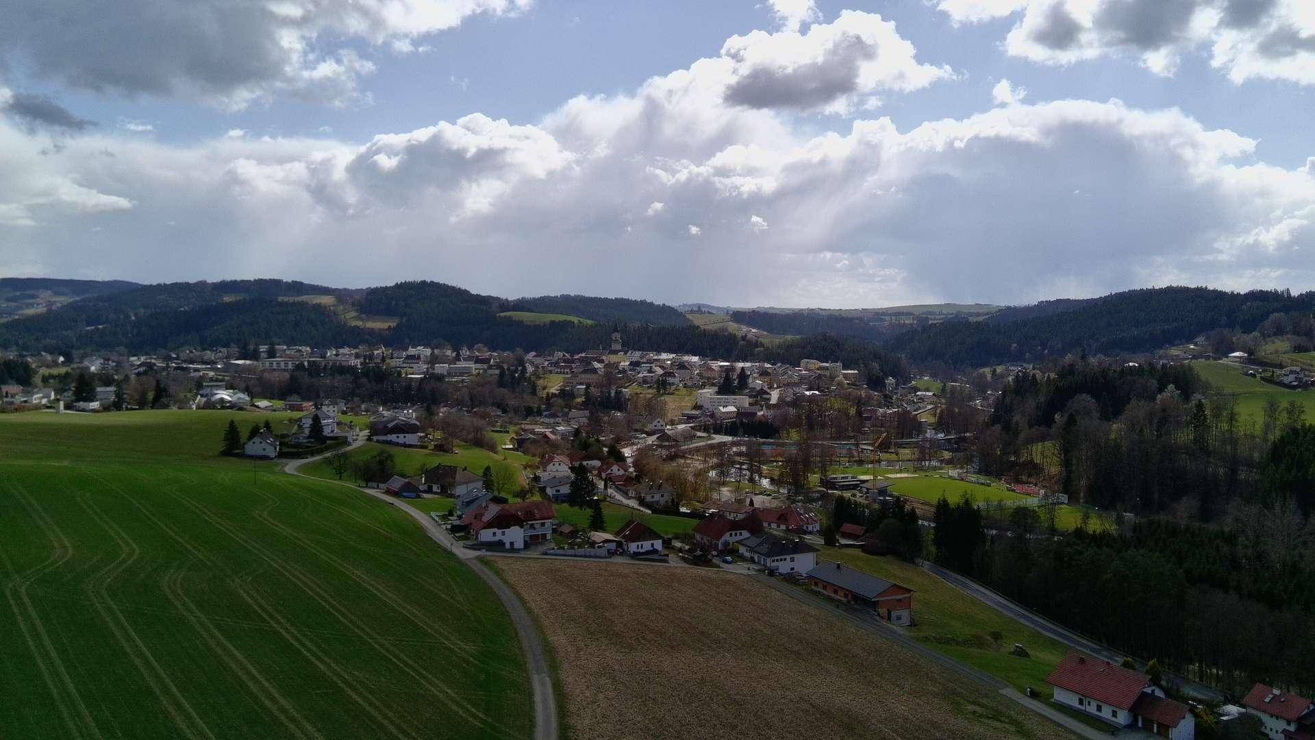 Luftaufnahme einer Kleinstadt in einer hügeligen Landschaft, mit Feldern im Vordergrund und bewölktem Himmel.