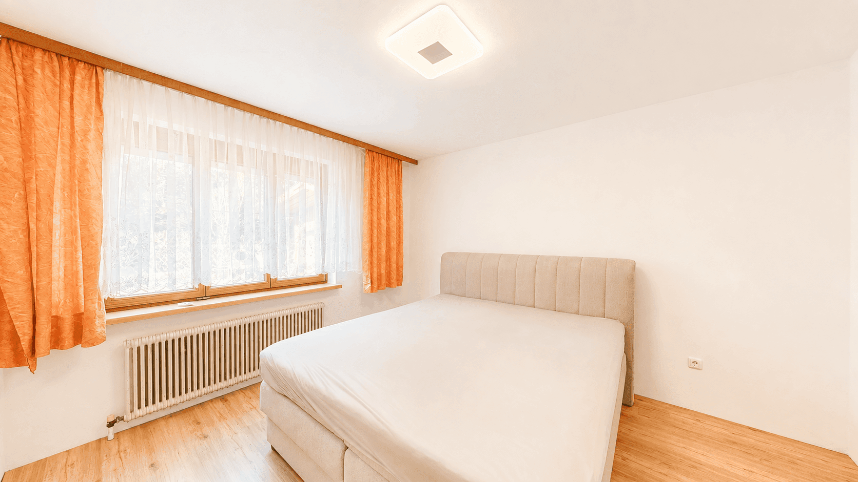 Helles Schlafzimmer mit Doppelbett, Laminatboden und einem Fenster mit orangefarbenen Vorhängen.